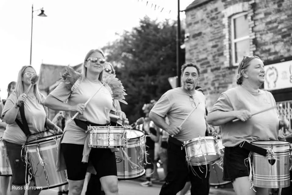 Group of people marching in a parade, playing drums with smiles and sunglasses, on a city street in black and white.