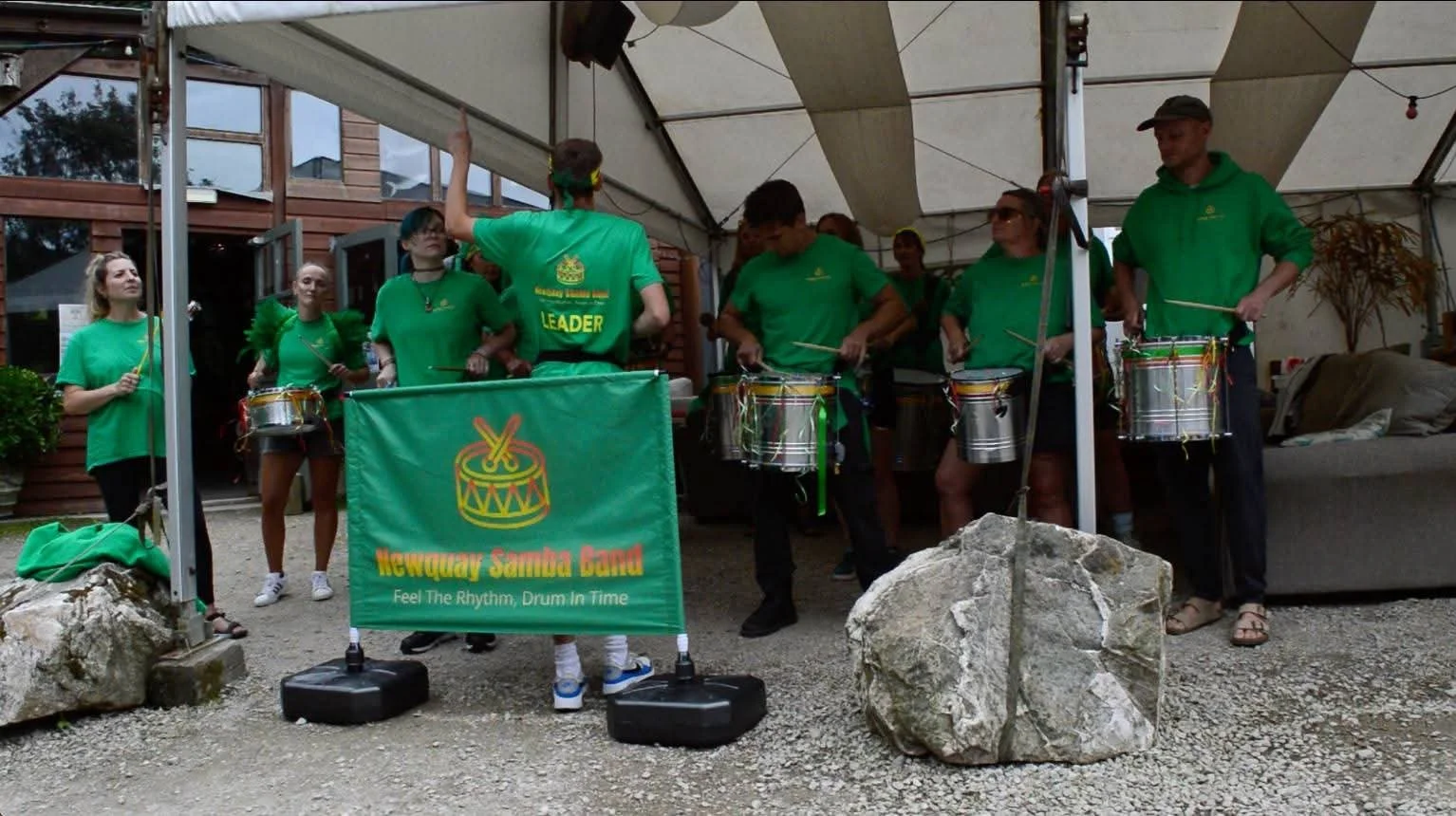 A group of people playing drums under a large outdoor tent, wearing matching green shirts with a banner that reads 'Newquay Samba Band' and 'Feel The Rhythm, Drum In Time'.