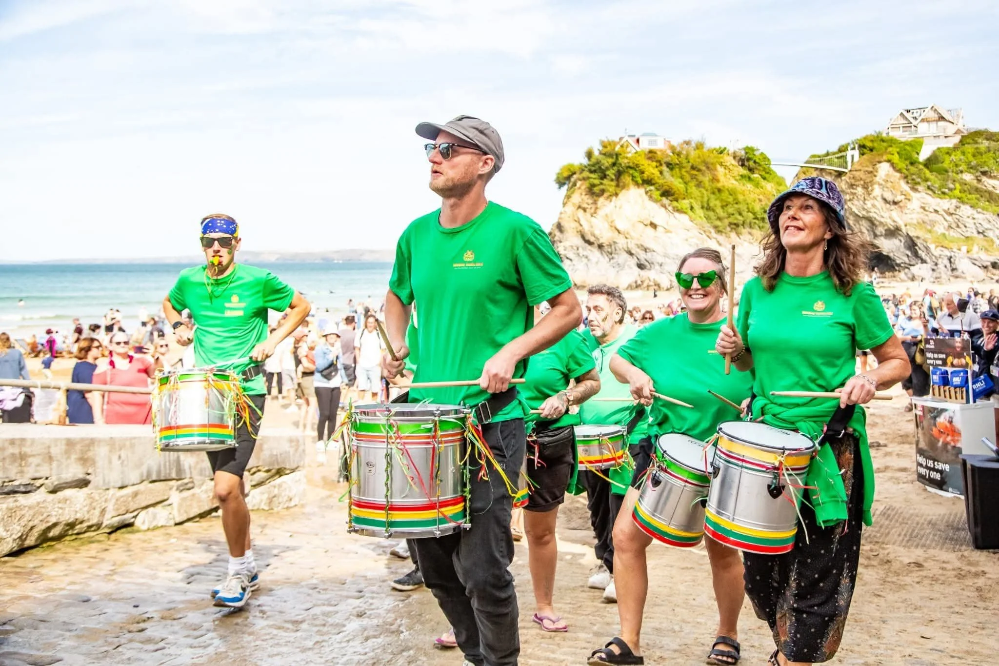 A group of people wearing green shirts, some with sunglasses or hats, marching on a beach with drums. There are many spectators in the background and a rocky hill with houses on top.