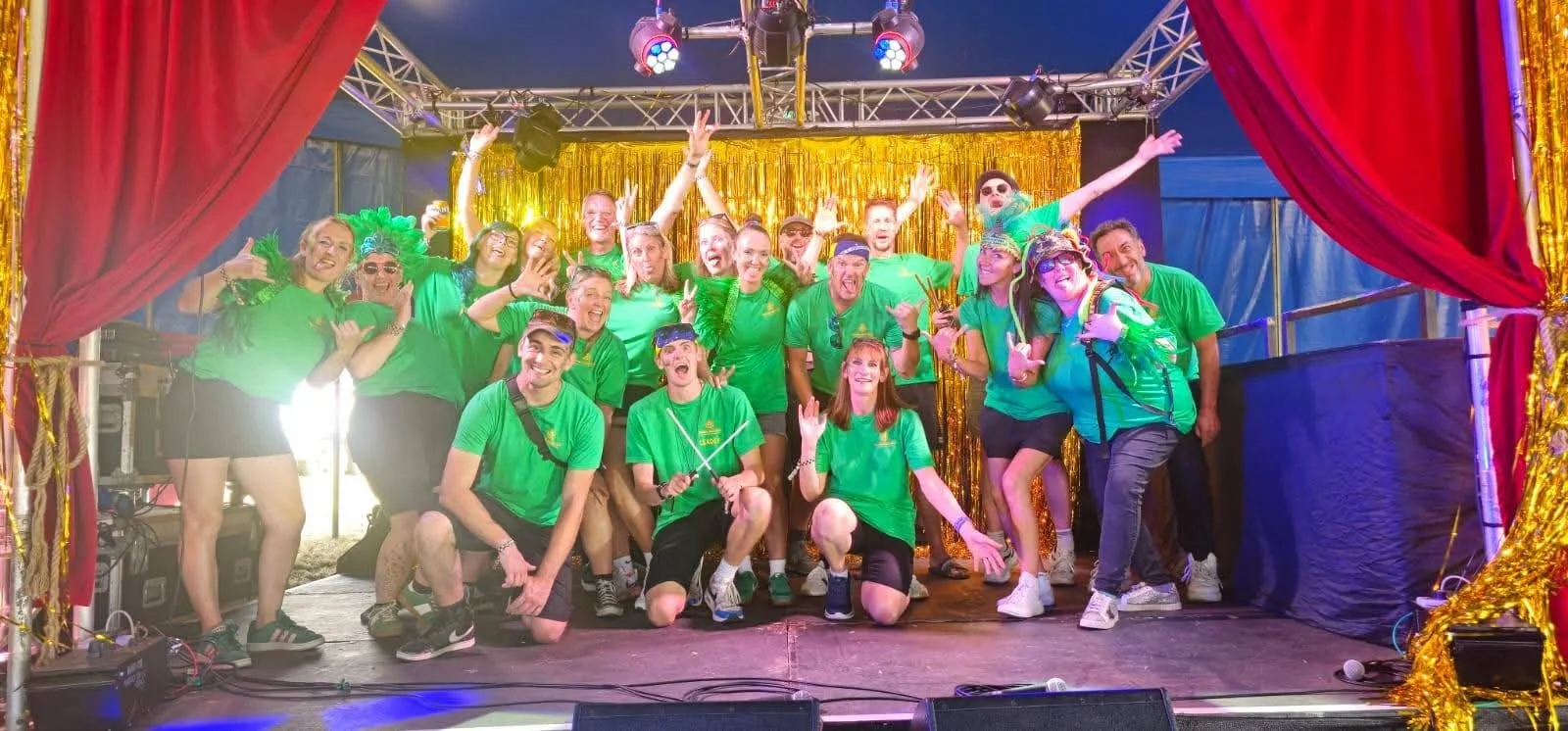 Group of people on a stage dressed in green shirts, some with accessories like bandanas and sunglasses, posing cheerfully with some making peace signs and others raising their hands, with red curtains and a gold backdrop behind them, indicating a cel