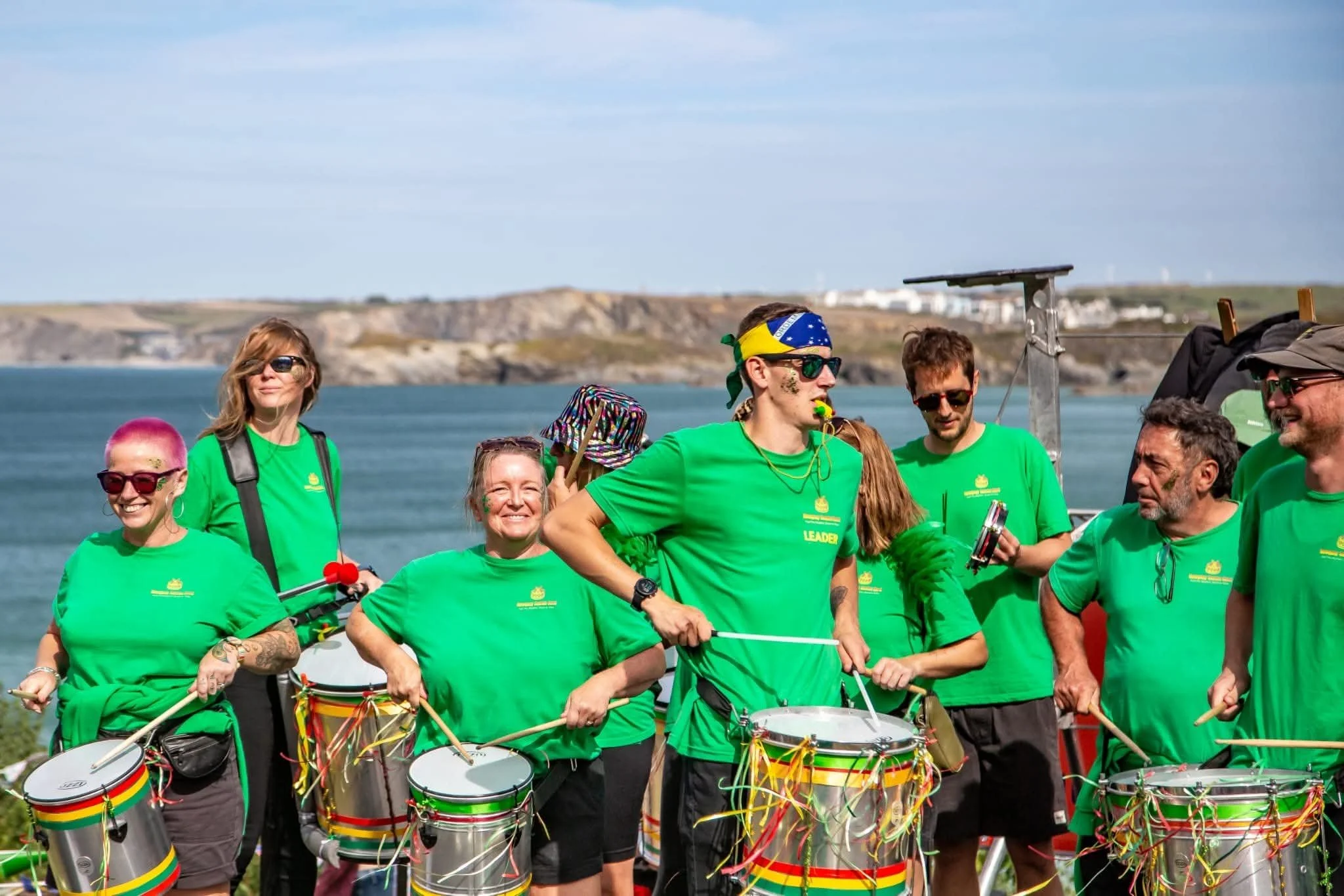 Group of people wearing green shirts, some holding drums, gathered outdoors near water with cliffs in the background.