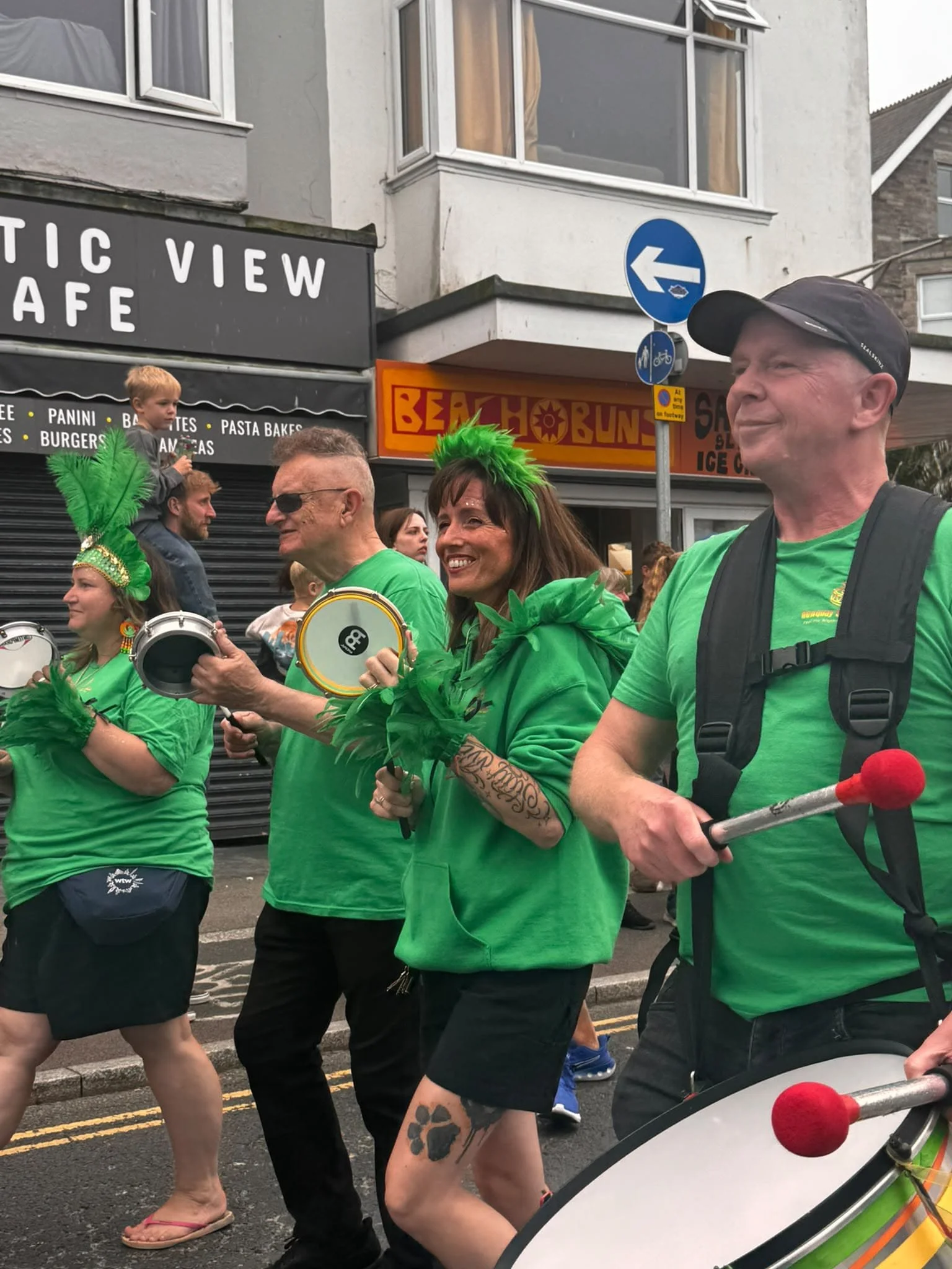 People walking in a parade, some wearing bright green shirts and feathered headpieces, with drums and other percussion instruments, on a city street with storefronts.