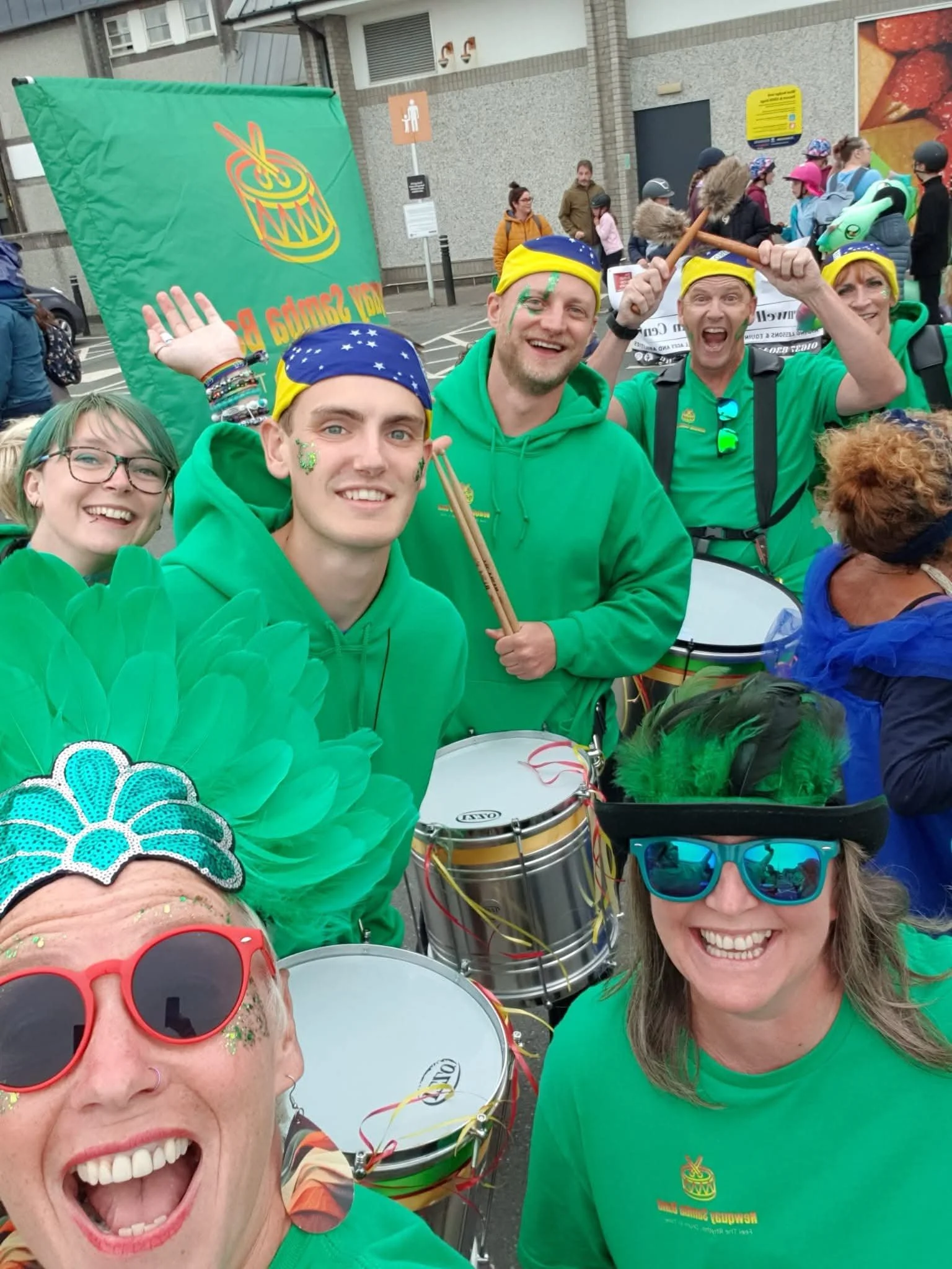 Group of people dressed in green shirts and colorful accessories smiling at a parade or festival, with drums and a green banner in the background.