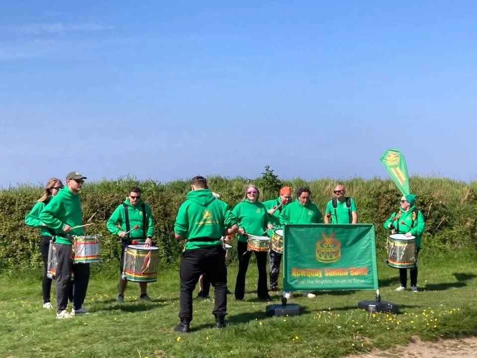 A group of people dressed in green hoodies standing outdoors on a grassy field, playing drums as part of a musical performance or parade.