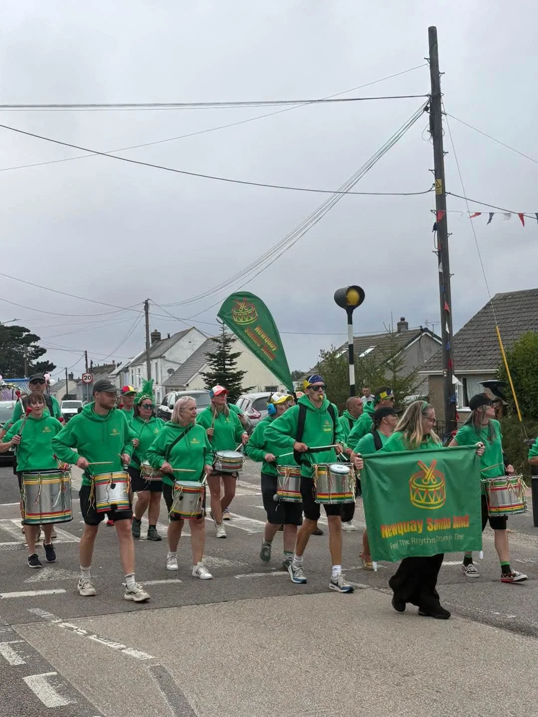 A parade of people wearing green hoodies and shorts, playing drums and carrying a banner that reads 'Newquay & St. Dennis' with a drum logo. The parade is on a street with houses and a cloudy sky overhead.