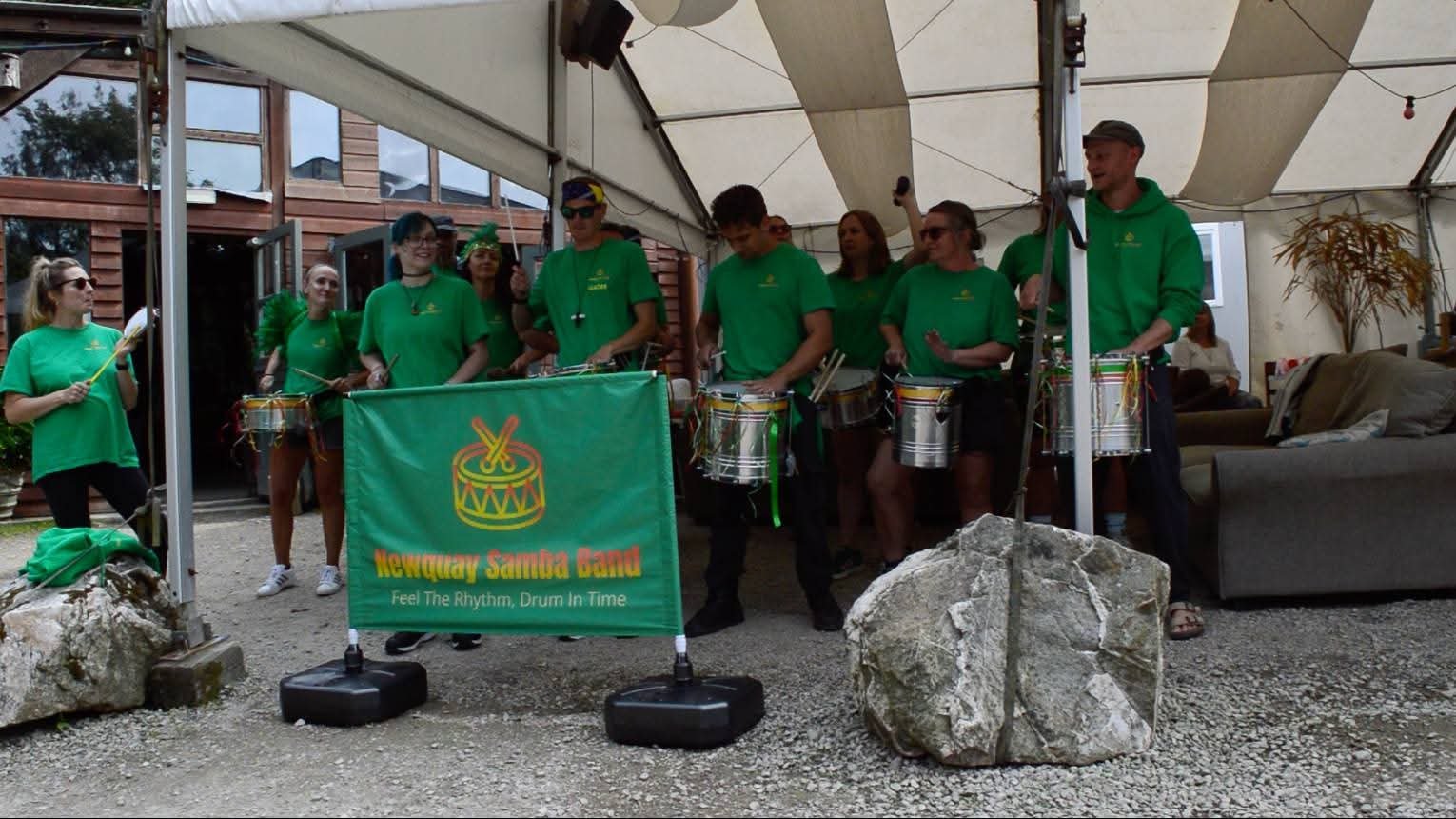 A group of people in green shirts playing drums under a tent, with a banner reading 'Newquay Samba Band' and 'Feel The Rhythm, Drum In Time.' They are outside near a wooden building and a sitting area with a couch.