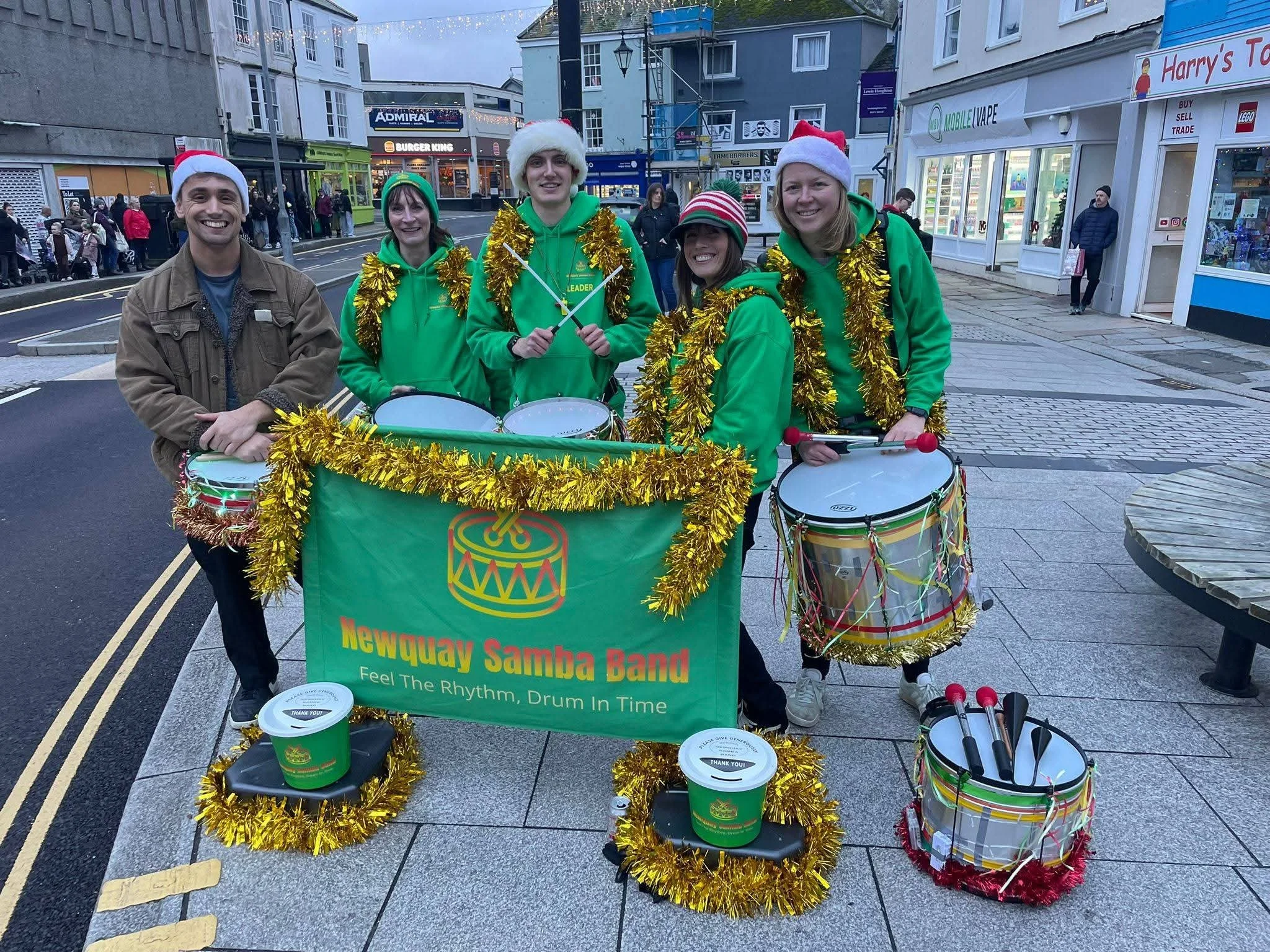 Group of five people wearing holiday hats and green sweatshirts, standing behind a drum set with yellow tinsel decorations, in a street with shops in the background. They are part of a band called 'Newquay Samba Band.'