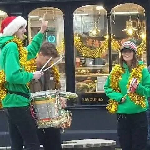 Three people celebrating Christmas outside a shop decorated with gold tinsel, with two women wearing green hoodies and Santa hats, one playing drums and the other holding a can, and a man in between wearing a grey hoodie.