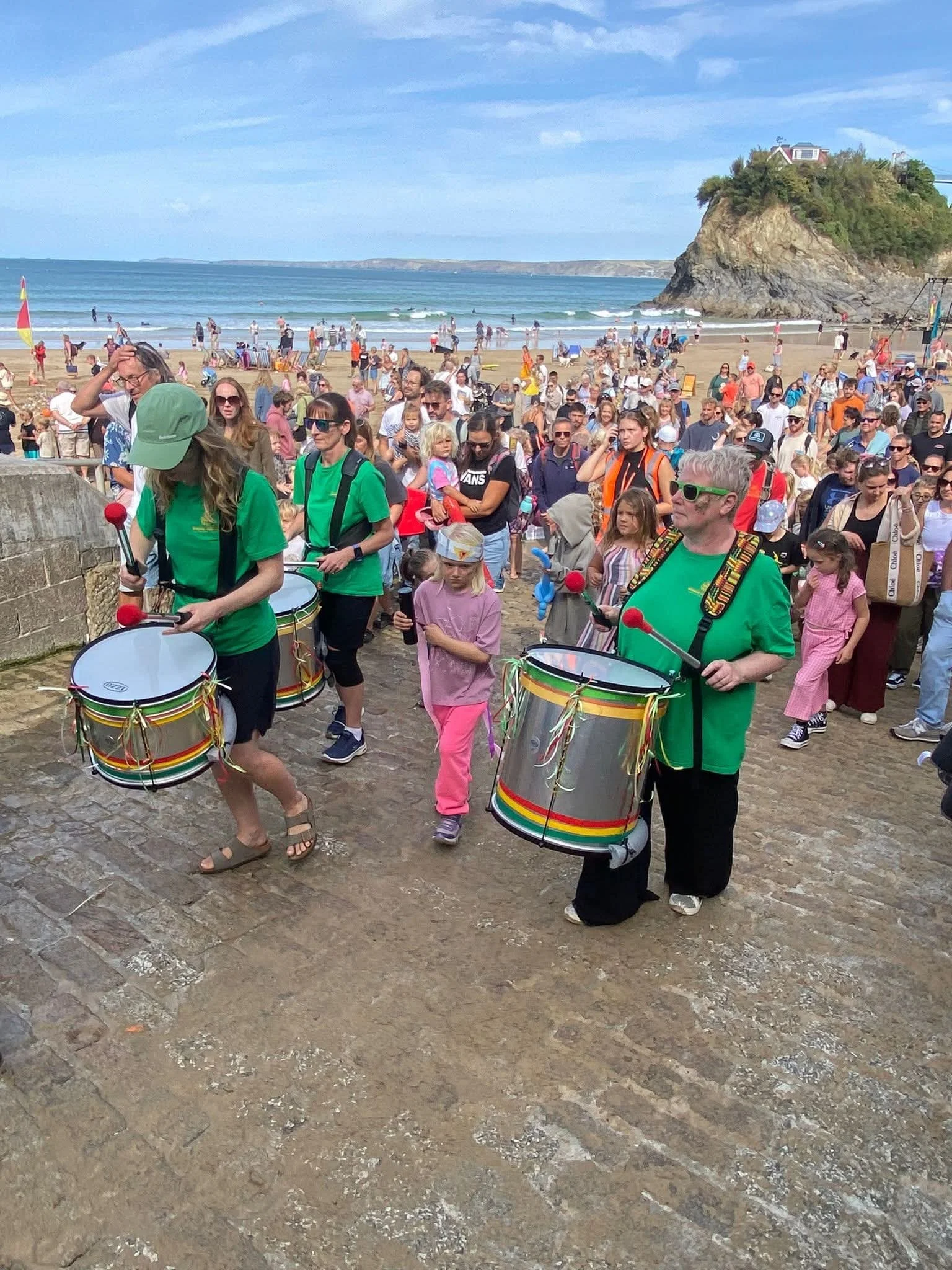 Crowd of people at the beach walking along the boardwalk, with some playing drums, near a rocky cliff with houses on top, under a partly cloudy sky.