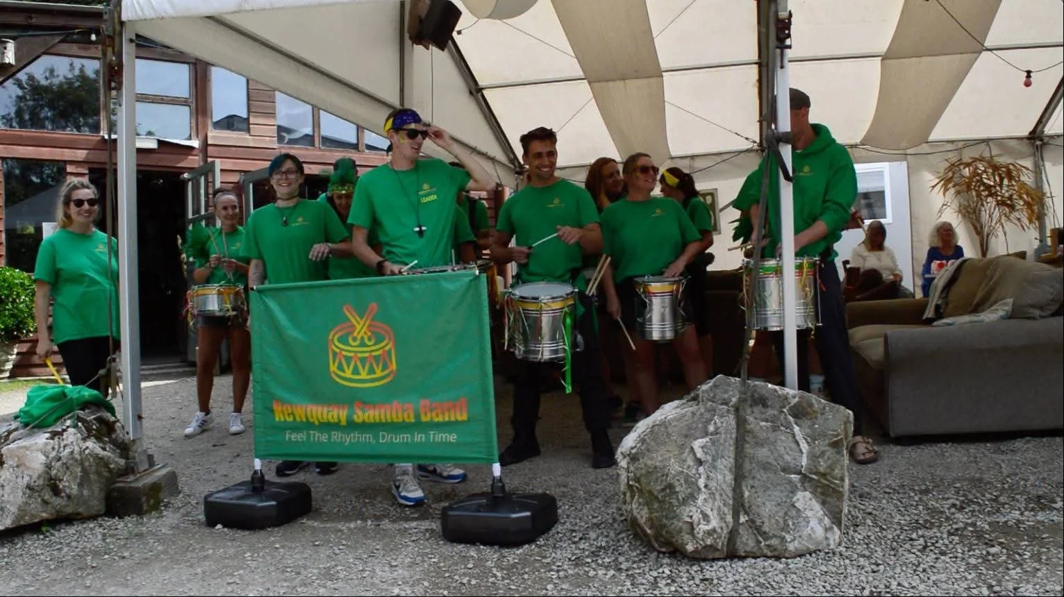 A group of musicians from the Newquay Samba Band performing under a canopy, playing drums and wearing matching green T-shirts.