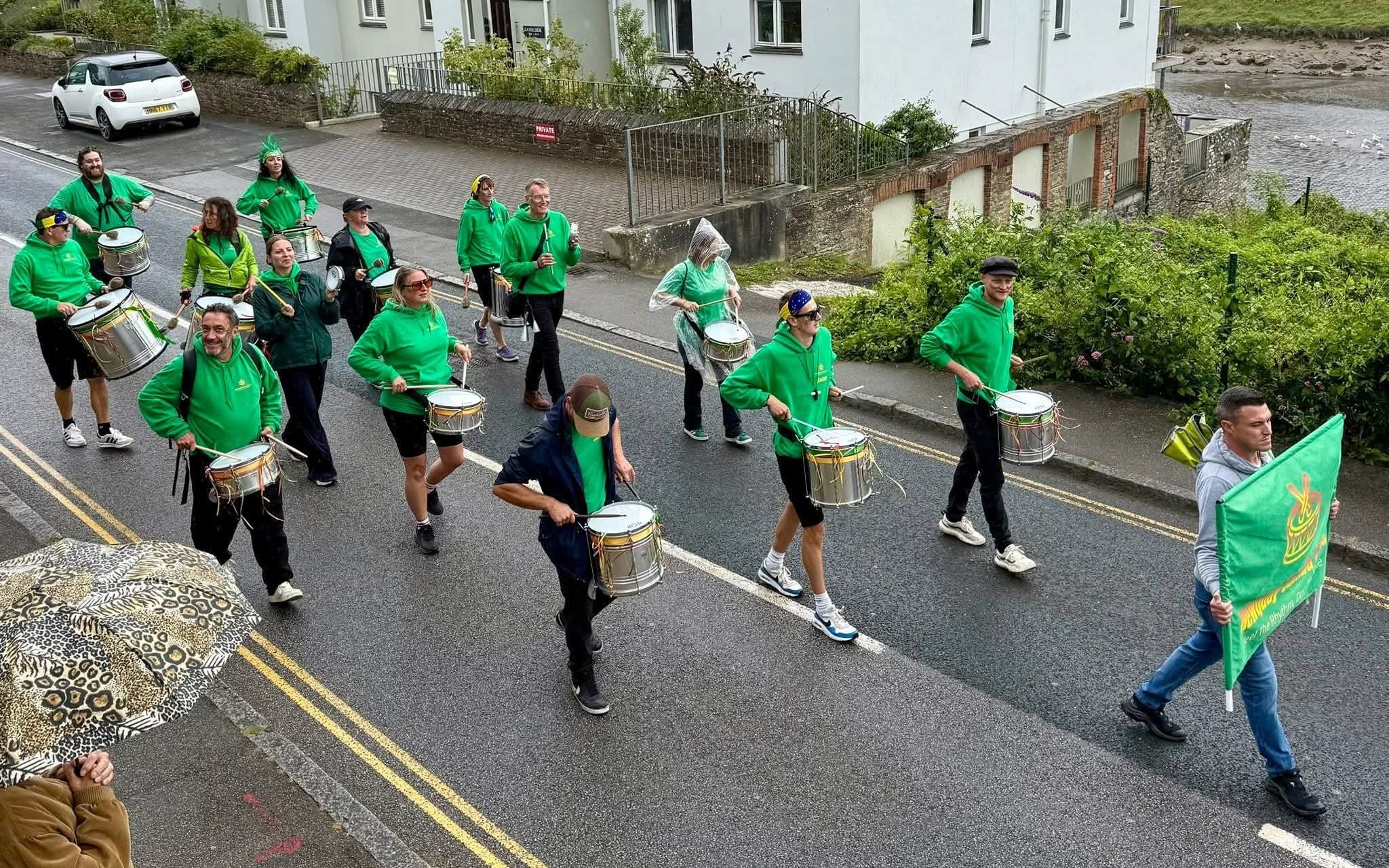 Group of people walking down a street playing drums, many wearing green hoodies, one holding a flag, some using umbrellas, with a building and parked cars in the background.