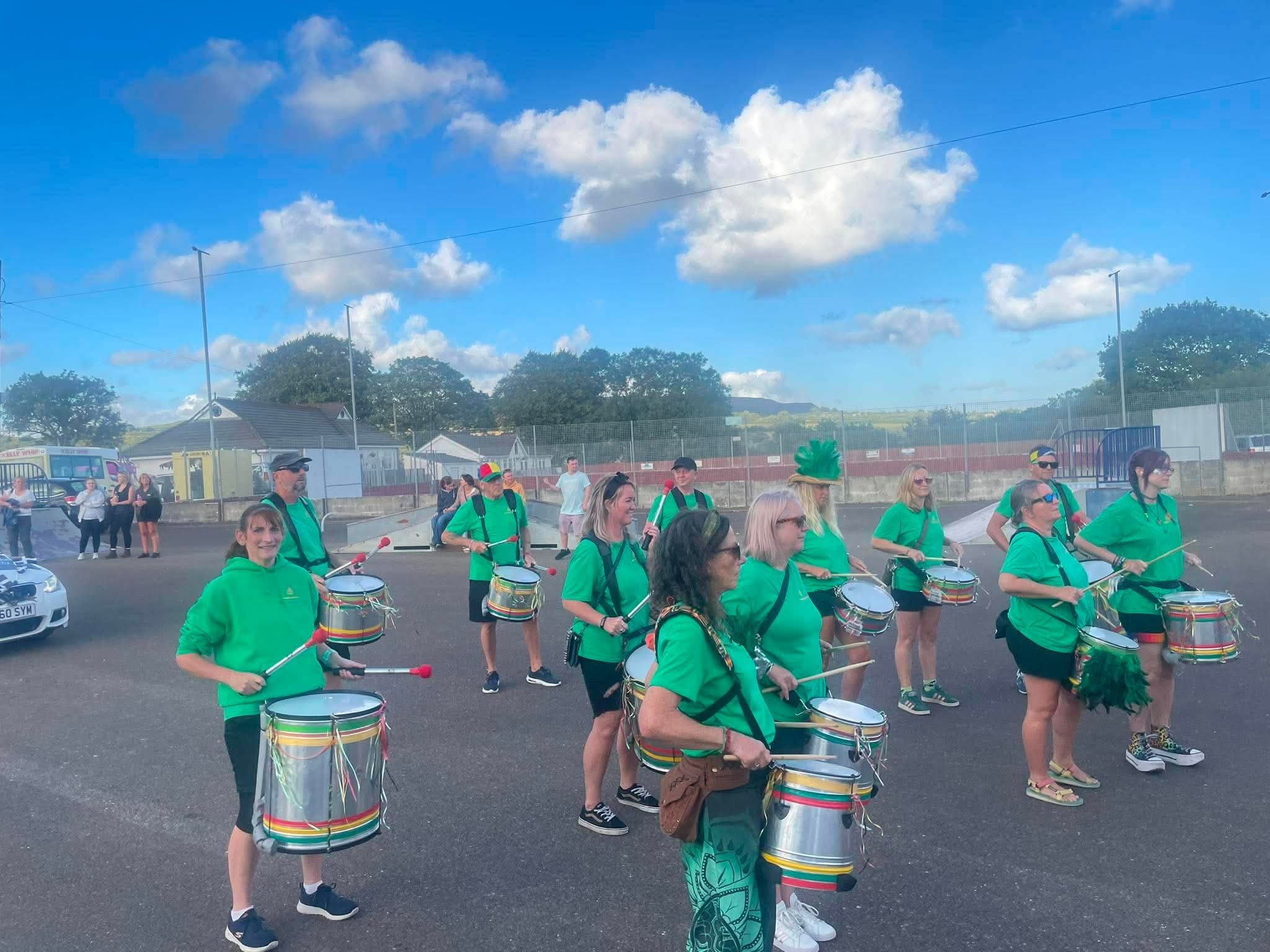 Group of people wearing green shirts and playing drums at an outdoor event on a sunny day with a bright blue sky and fluffy clouds.