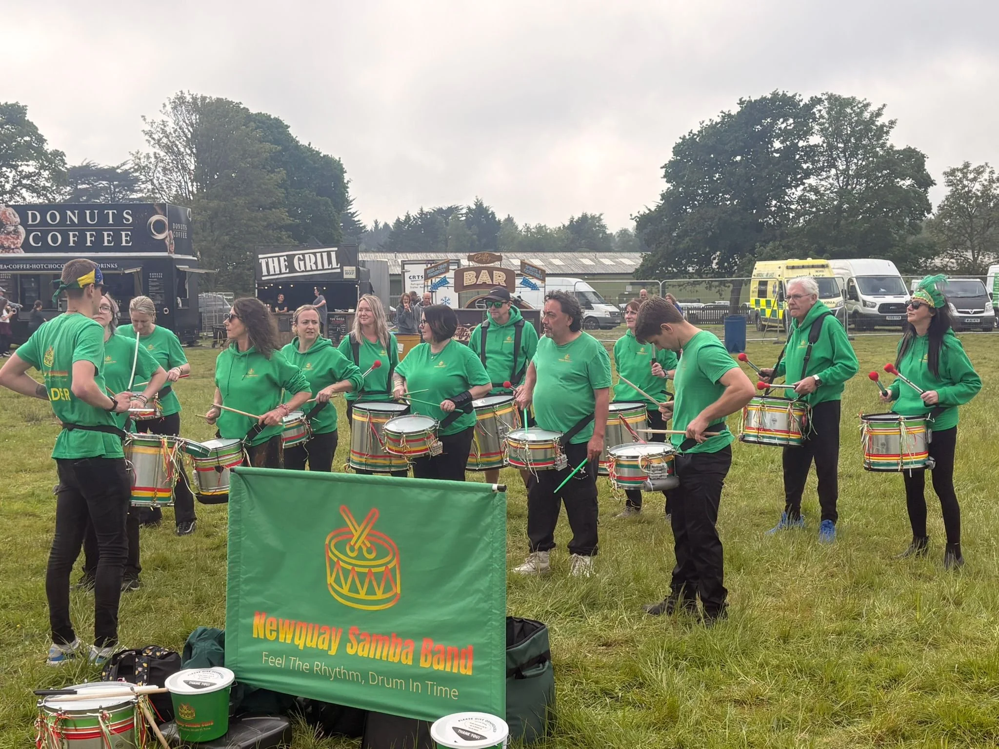 A group of people wearing green shirts, some with hats or sunglasses, are gathered outdoors playing drums in a samba band at a festival or event. Behind them, there is a food truck offering donuts and coffee, a bar, and other event signage. Trees and