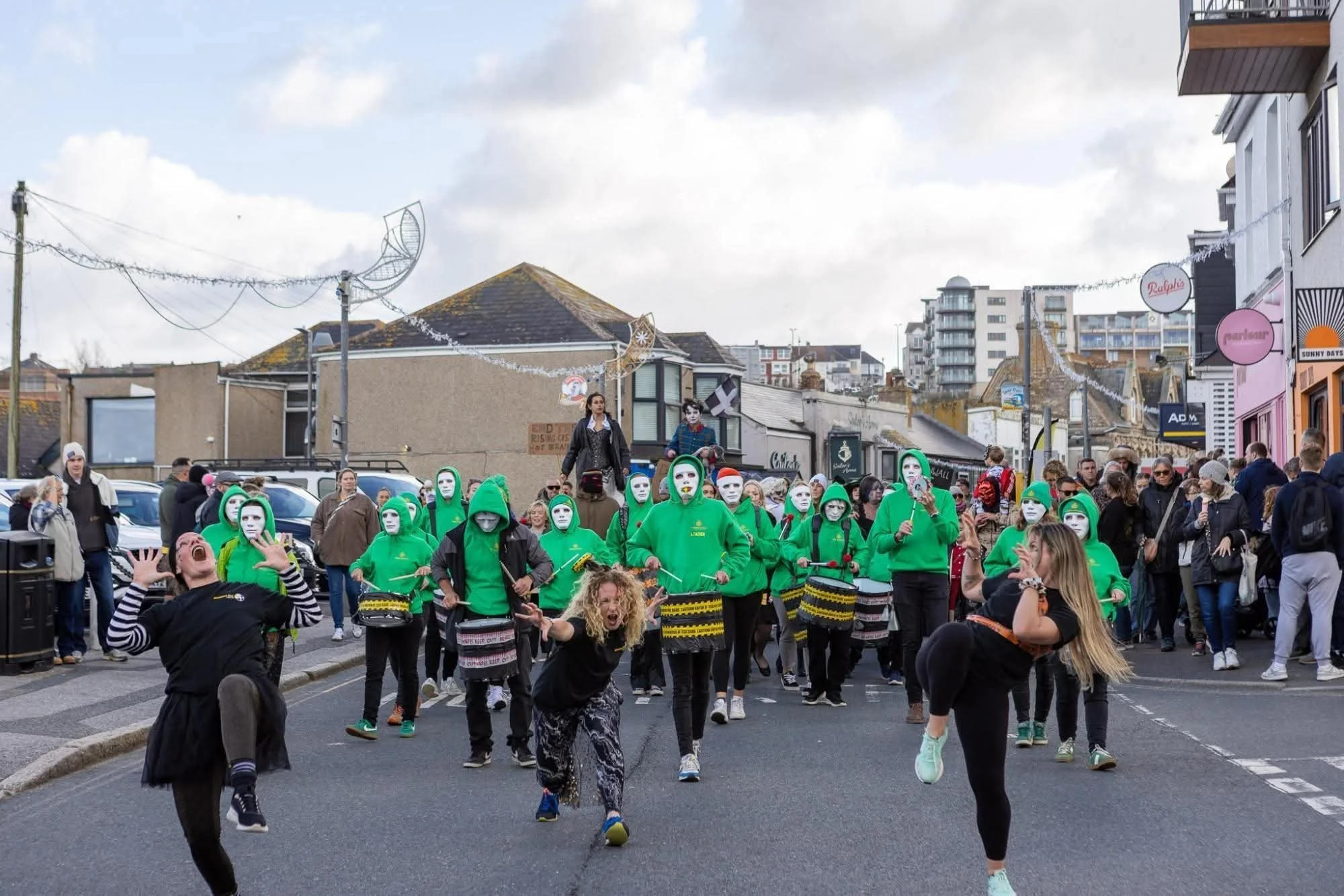 A street parade with people wearing green and white masks, some playing drums, and a crowd watching. The background shows buildings and a cloudy sky.