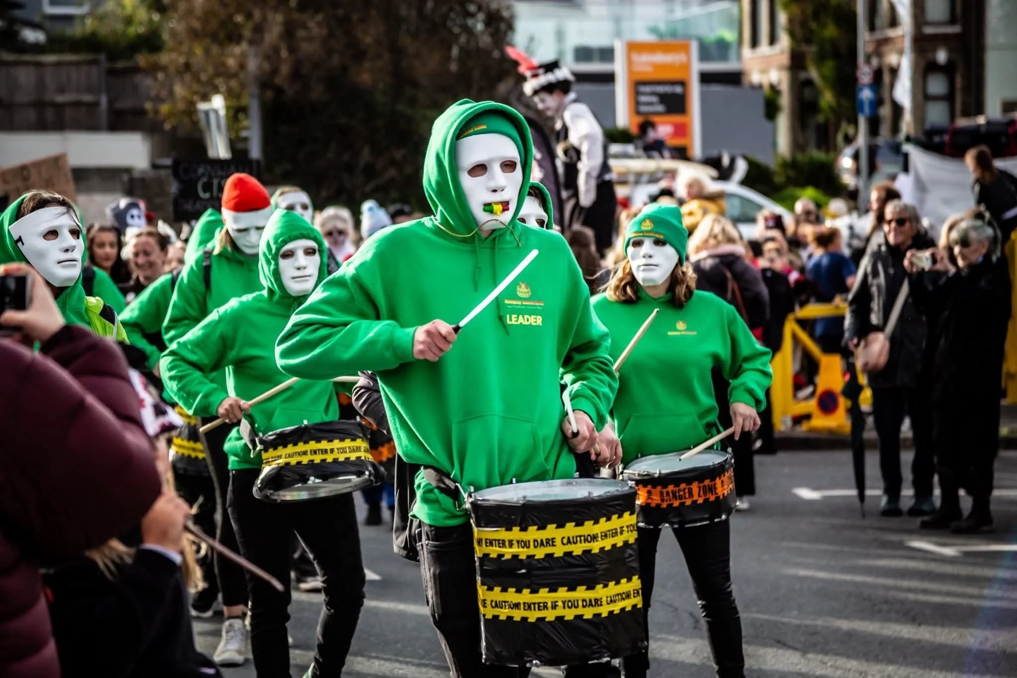 A group of people dressed in green hoodies and white masks participating in a parade or demonstration, with some wearing Halloween hats and holding drumsticks, marching on a street lined with spectators.