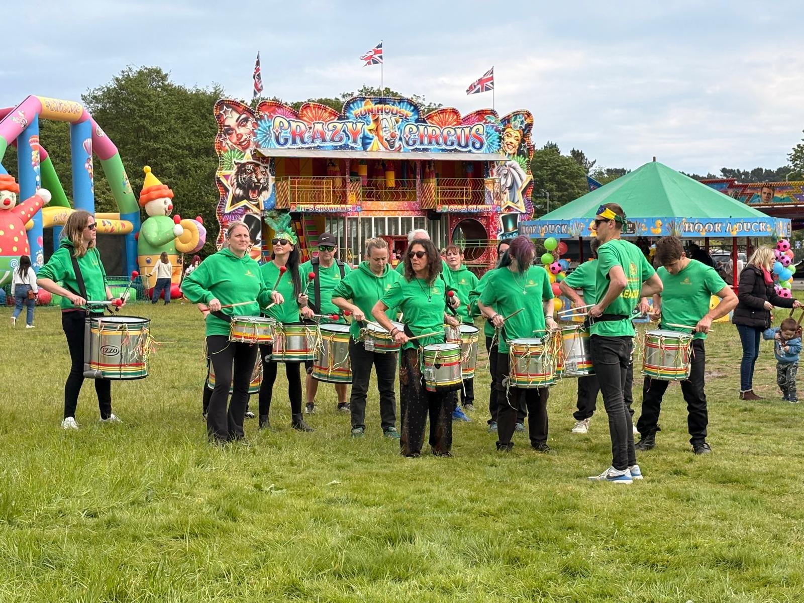People in green shirts playing drums at a carnival with colorful rides and attractions in the background.