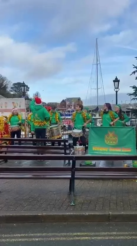 Group of people dressed in festive green and yellow costumes, some wearing Santa hats, standing near water with boats and harbor in the background, during a parade or celebration.