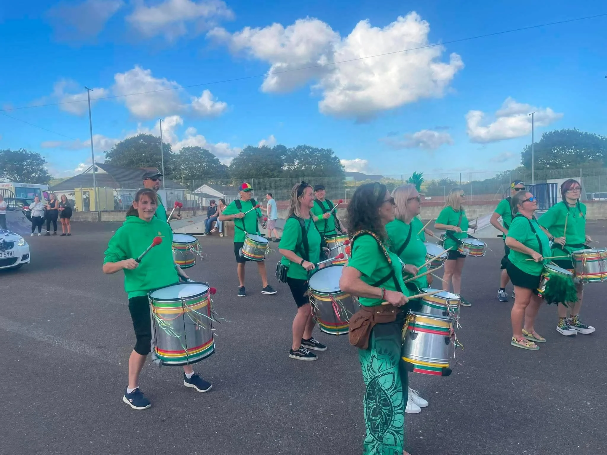 A group of people dressed in green shirts are marching, playing drums with colorful stripes, in an outdoor area. The background shows trees, buildings, a blue sky with scattered clouds, and a few onlookers.