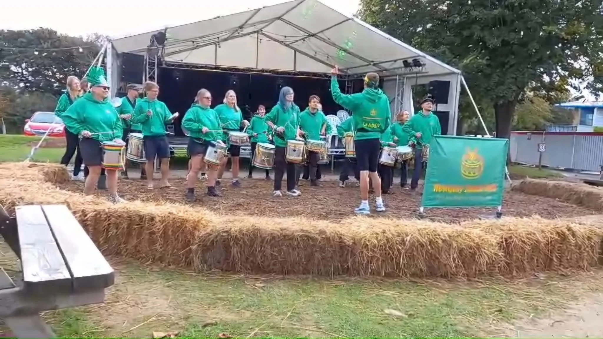 A group of people wearing green hoodies and shorts are performing on an outdoor stage. They are playing drums and are led by a conductor. The stage is decorated with hay bales and has a large tent overhead. Trees and parked cars are visible in the ba
