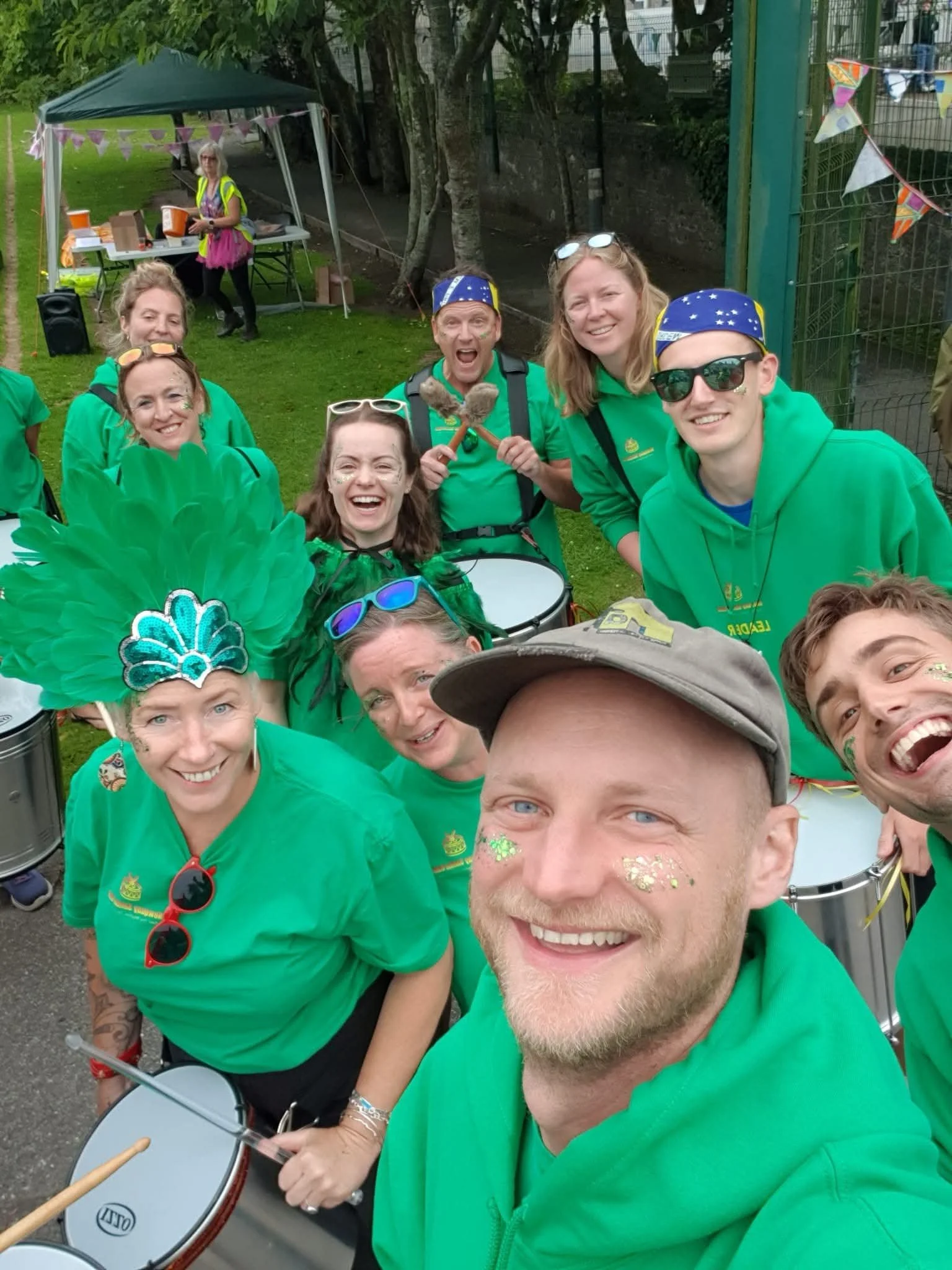 Group of people wearing green shirts, some with glitter face paint, celebrating outdoors with drums, fireworks, and festive accessories, possibly at a parade or festival.