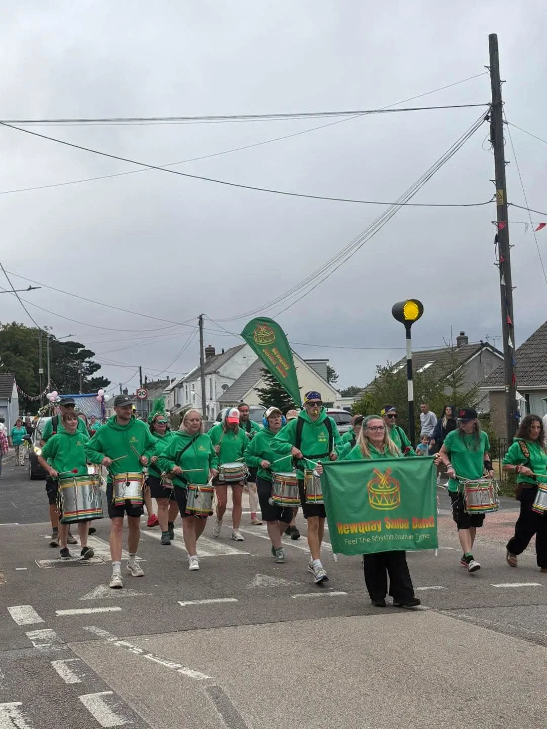 A parade with members of the Newquay Samba Band marching down a street, wearing green hoodies and playing drums, with houses and power lines in the background on a cloudy day.
