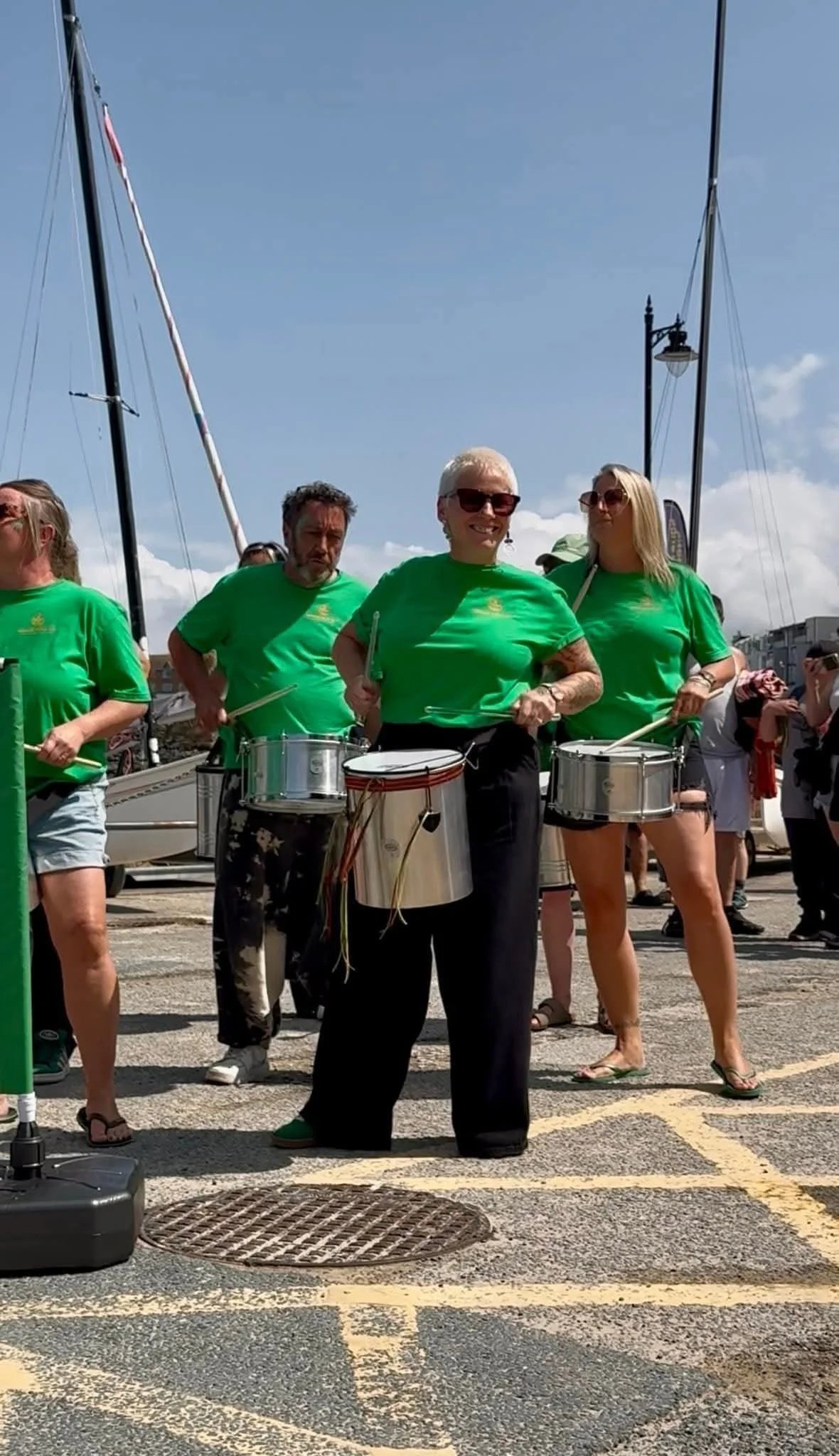 A group of people wearing green T-shirts playing drums outdoors on a sunny day, near sailboats and a marina.
