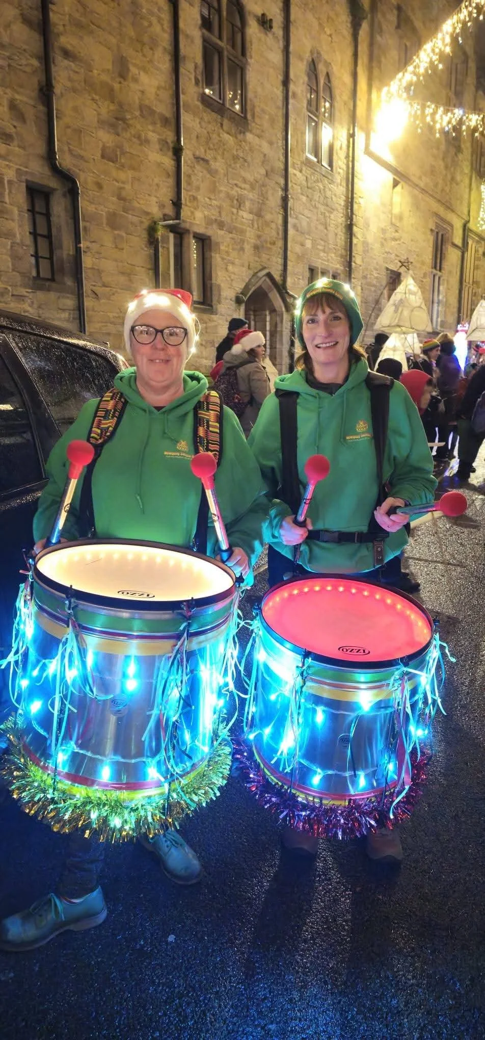 Two women dressed in green hoodies and Santa hats, holding drumsticks, and standing behind illuminated drums decorated with Christmas lights at a festive outdoor event at night.