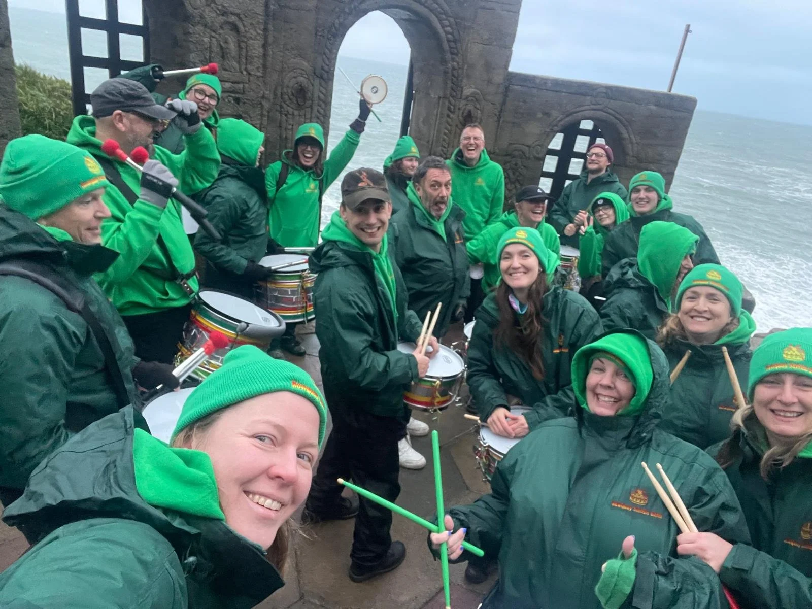 A group of people dressed in green jackets, hats, and gloves are gathered on a ship. They are smiling and playing drums and percussion instruments. The background shows a cloudy sky and ocean, with the ship's structure and an archway.