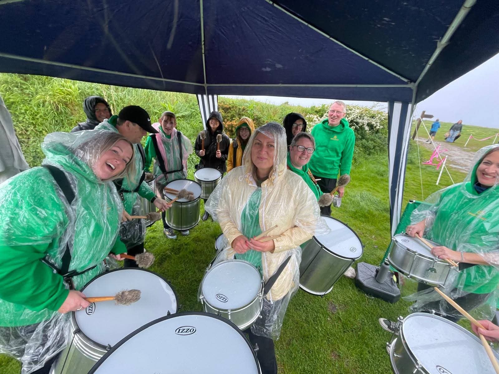 Group of people playing drums under a tent outdoors on a rainy day, wearing raincoats and holding drumsticks, with greenery and other people in the background.