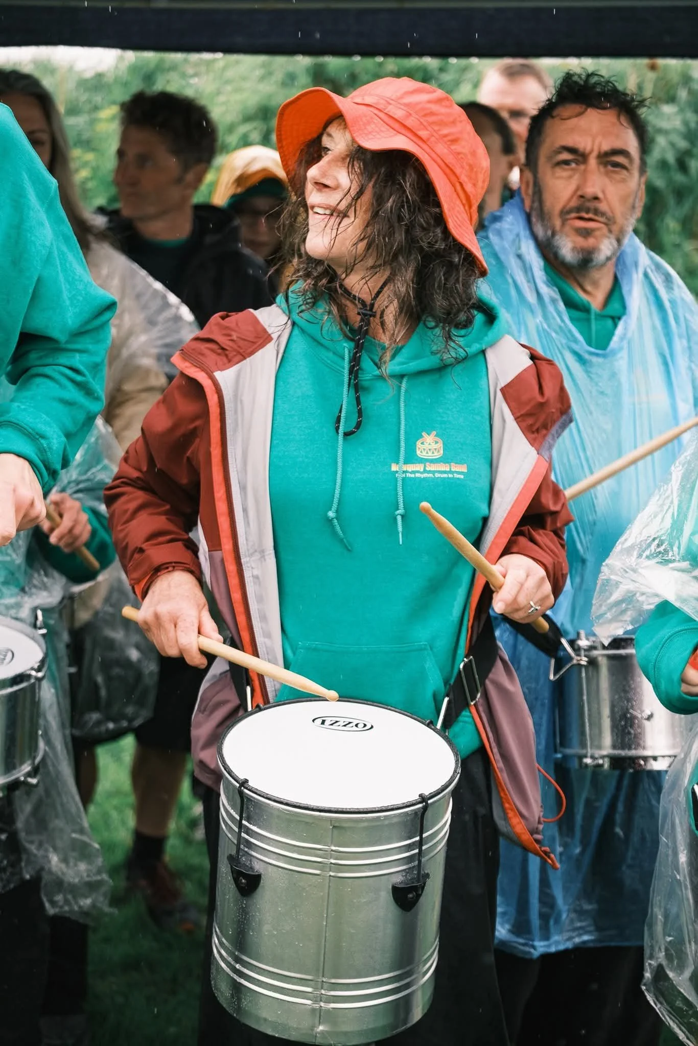 A group of people playing drums outdoors, with a woman in the foreground wearing a red hat and teal shirt, holding drumsticks, and smiling. Other individuals are visible in the background, some holding drumsticks as well, with greenery in the backgro