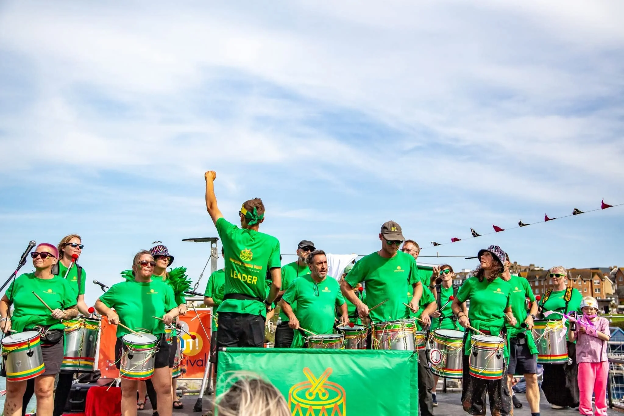 A group of people wearing bright green shirts participating in a public event, with some holding drums, under a partly cloudy sky.