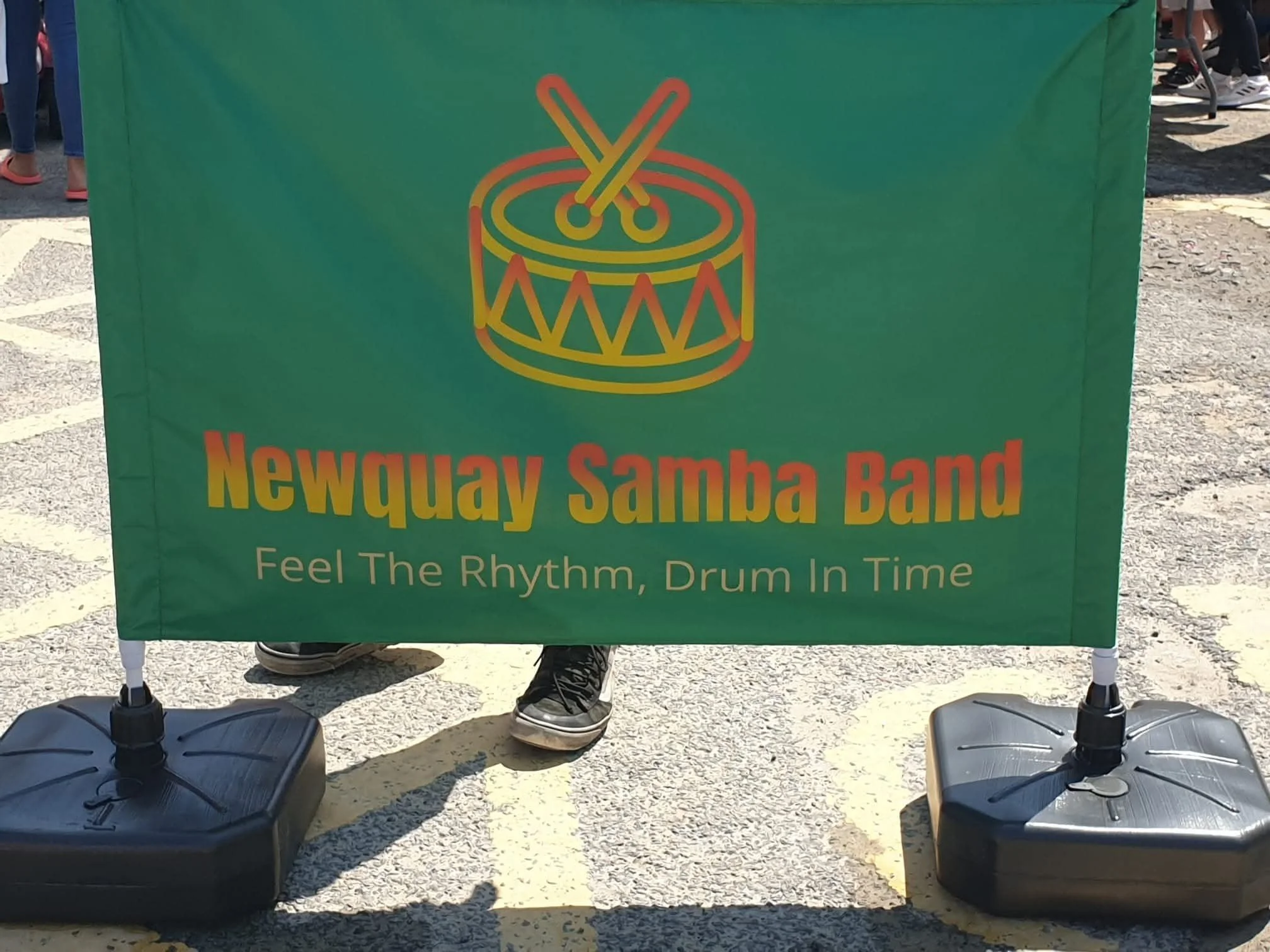 A green banner with a colorful neon-style drum and drumsticks graphic and text that reads "Newquay Samba Band" and a tagline to feel the rhythm and drum in time. The banner is held on stands, and people are visible in the background at an outdoor eve