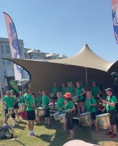 A group of people wearing green shirts playing drums and percussion instruments under a large tan canopy tent outdoors, with residential buildings and blue sky in the background.