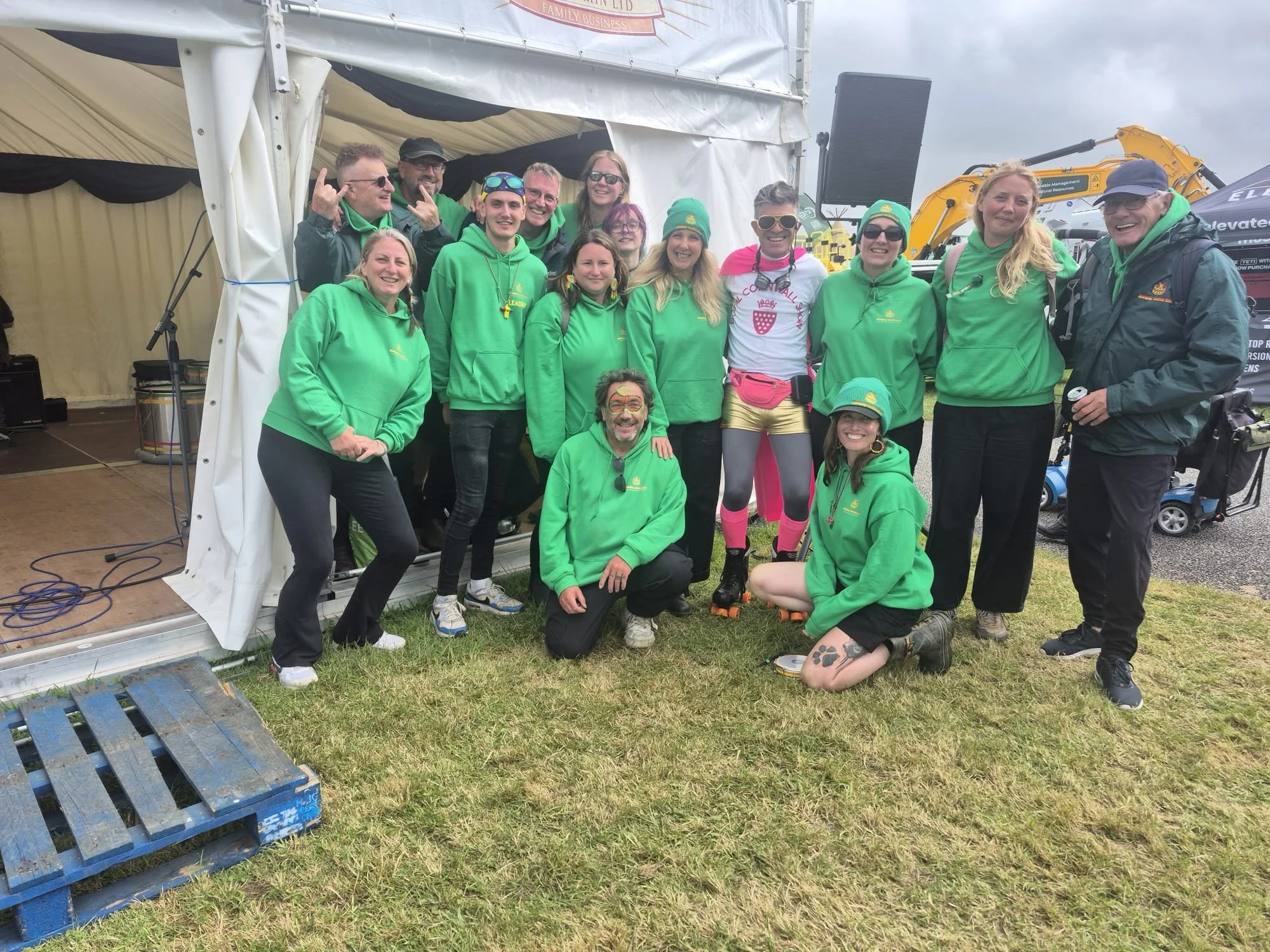 Group of people posing for a photo at an outdoor event, some wearing green hoodies and others in colorful athletic outfits, standing in front of a white tent and construction equipment.