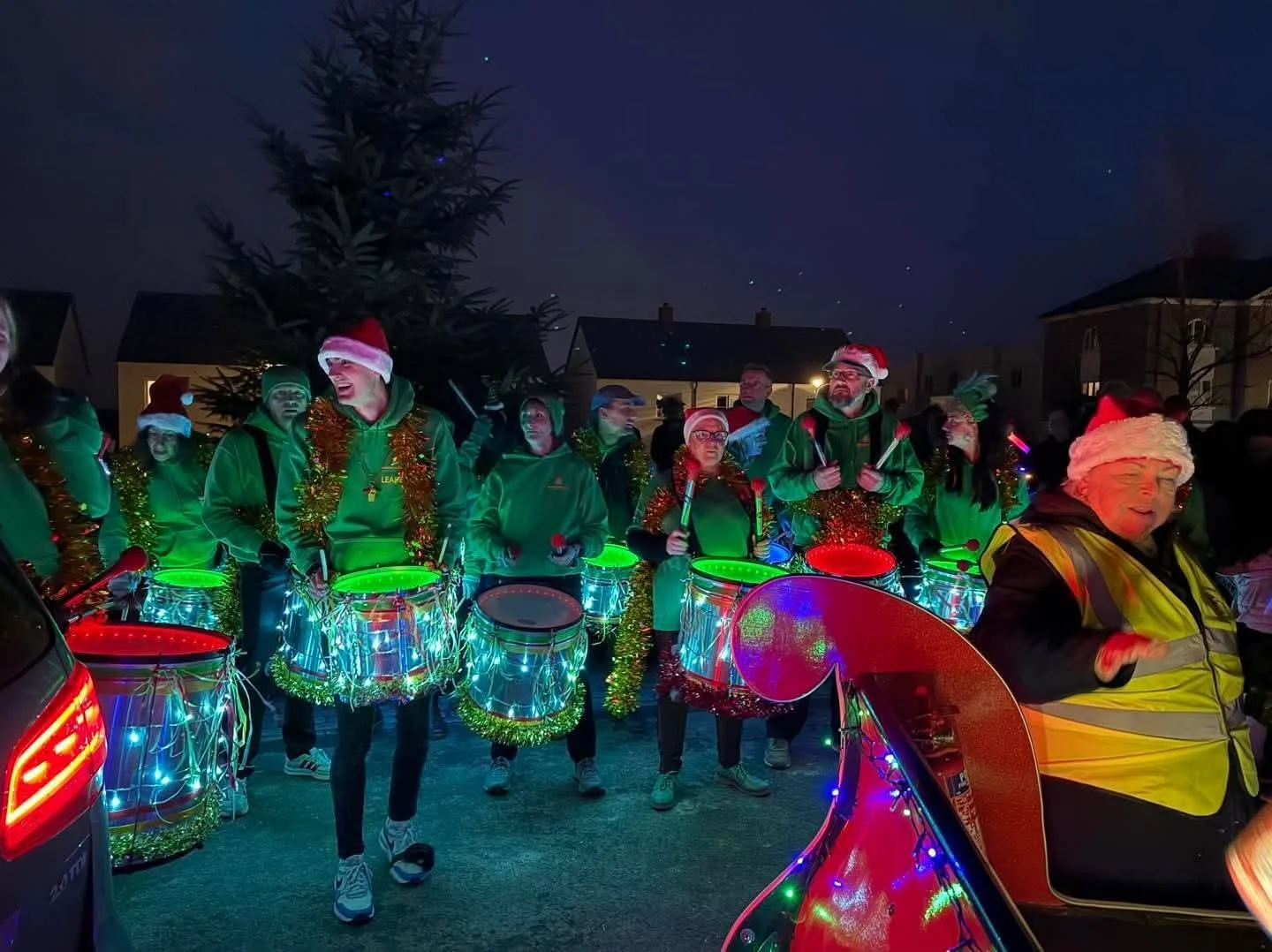 Group of people dressed in Christmas attire playing drums decorated with Christmas lights during a night parade, with houses and a large Christmas tree in the background.