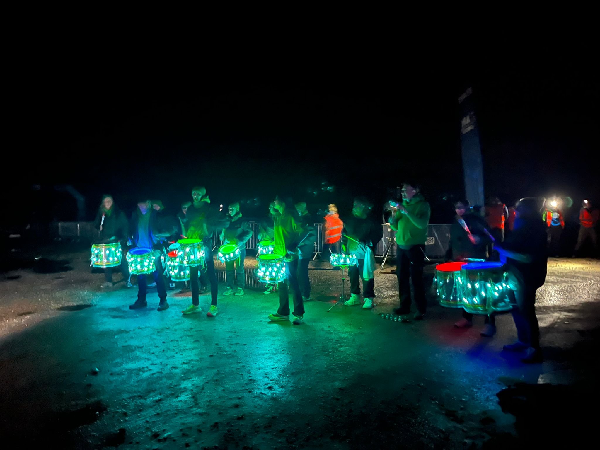 Night scene of a group of people gathered outdoors, illuminated by glowing drums and some wearing reflective vests, with a dark sky and a tall structure in the background.