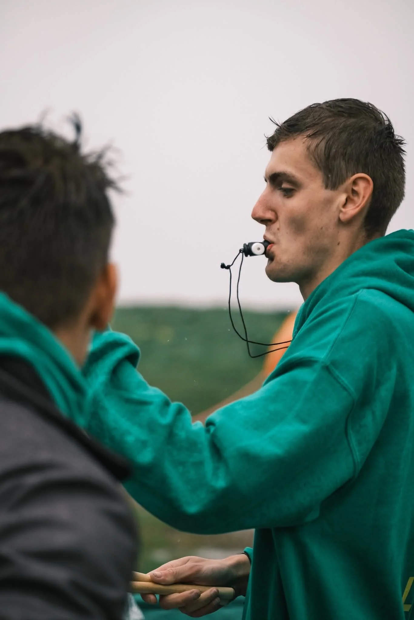 Two young men are outdoors, engaging in a handshake while one is handing the other a wooden object. Both are wearing teal hoodies.