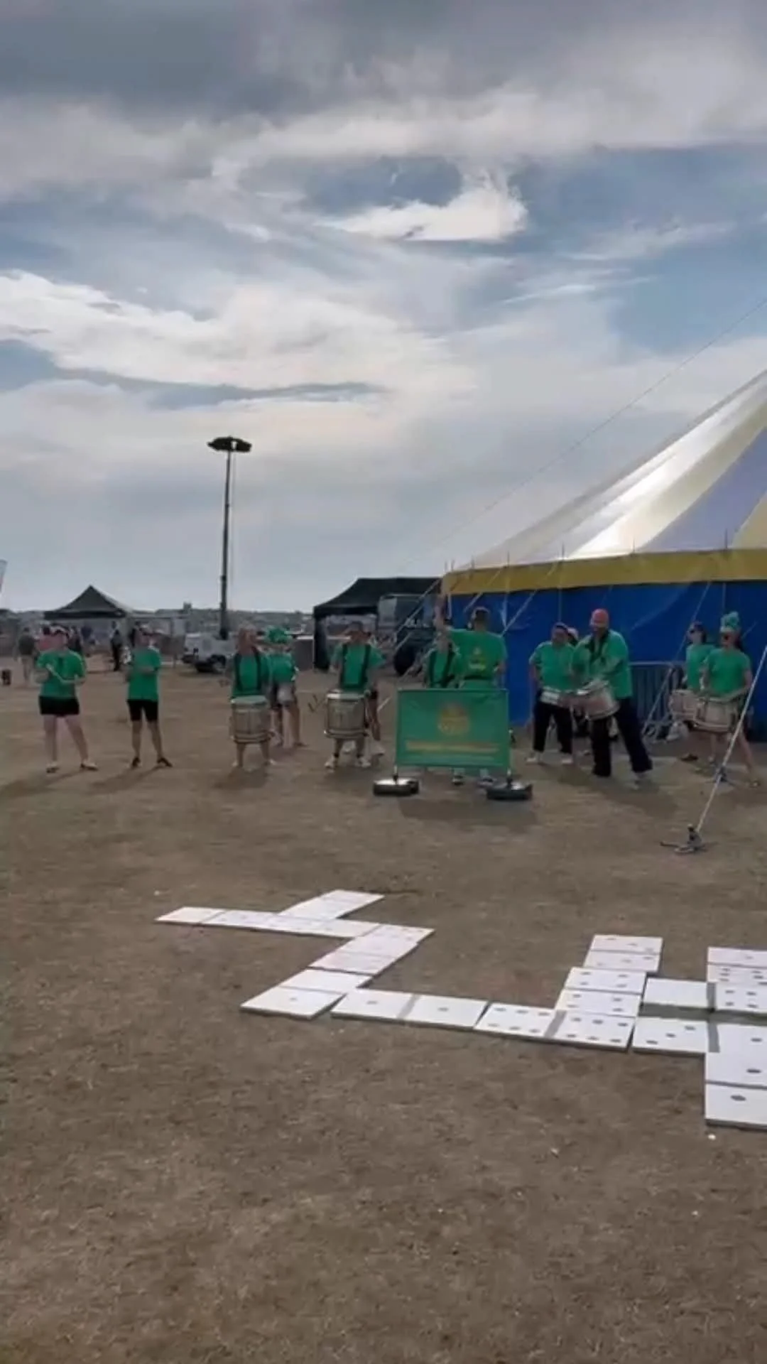 Music performers playing drums at an outdoor event with tents and people in the background under a cloudy sky.
