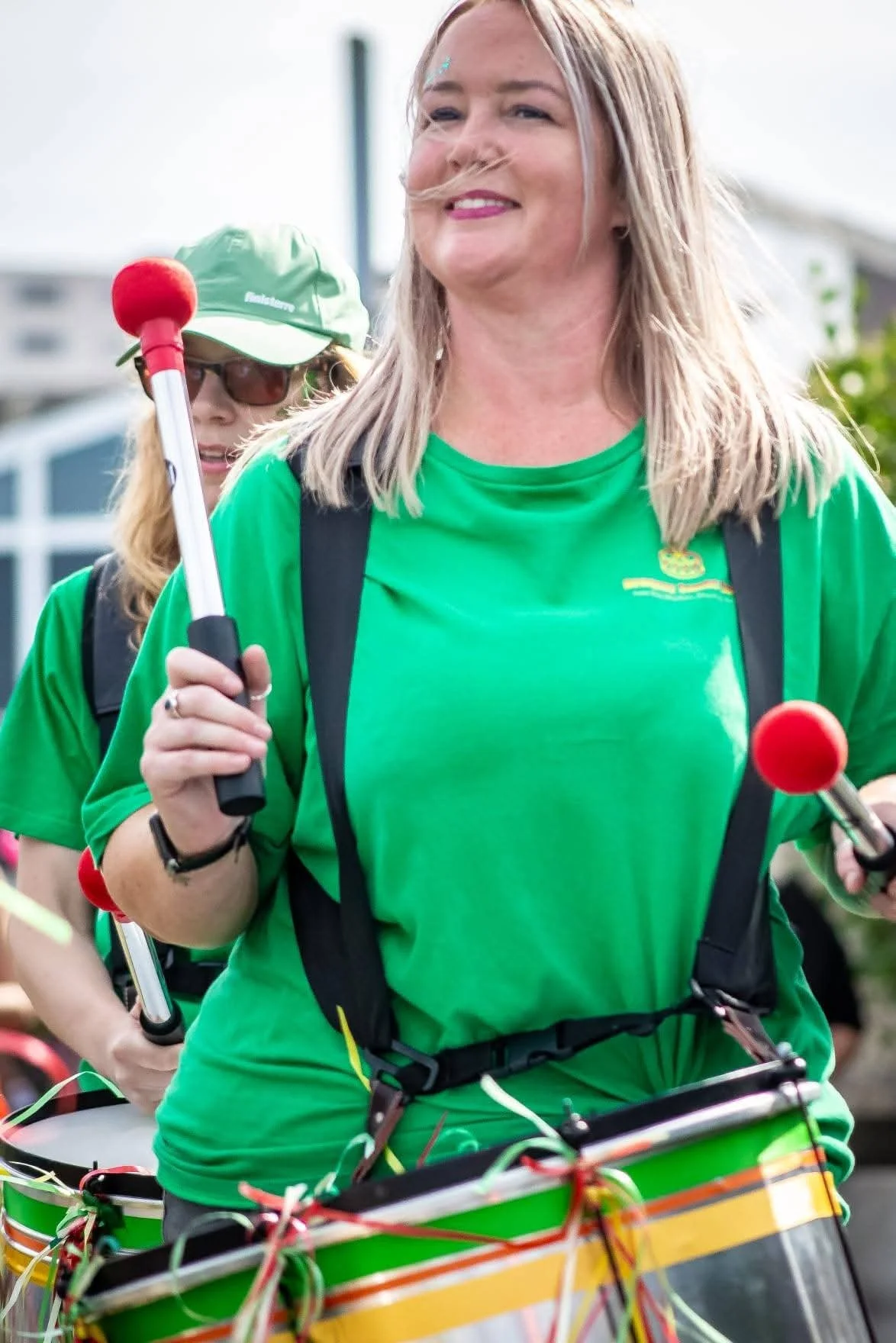 A woman smiling while riding a decorated bicycle. She is wearing a green shirt and a backpack, with another woman in the background also wearing a green hat and sunglasses.