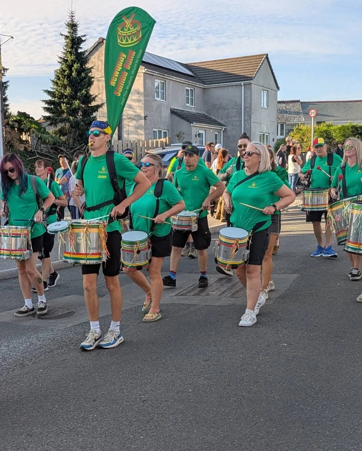 A group of people wearing green shirts and playing drums march on a street during daylight. Some participants wear sunglasses and backpacks, with one individual leading. In the background, there is a house, a 20 km/h speed limit sign, and a lamppost,