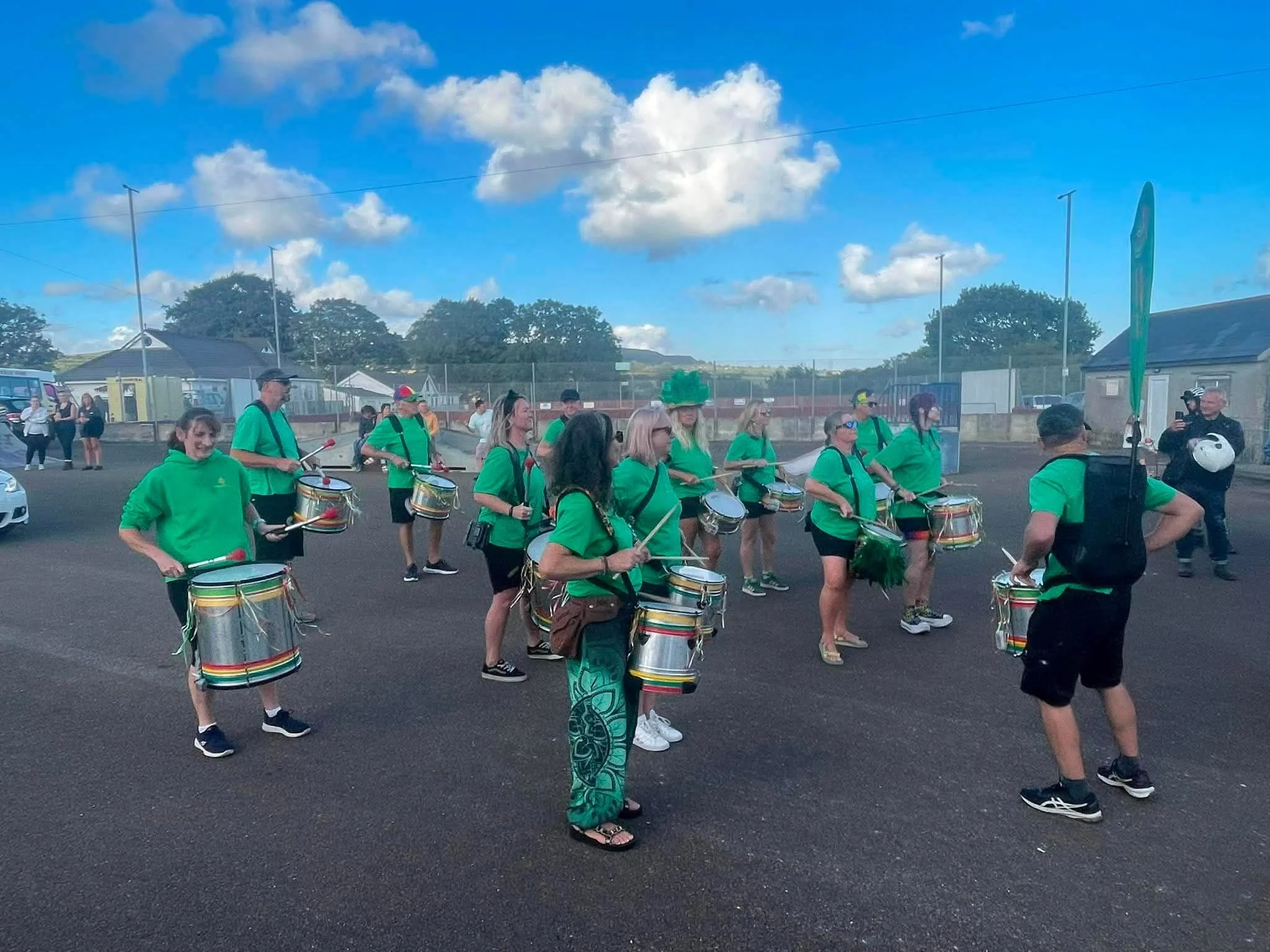 A group of parade musicians dressed in green shirts playing drums in an outdoor parking lot under partly cloudy sky.