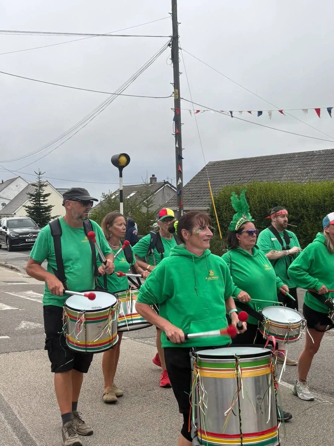 A group of people, mostly women, in green shirts and hoodies, playing drums and marching in a parade on a street with houses and cars in the background.