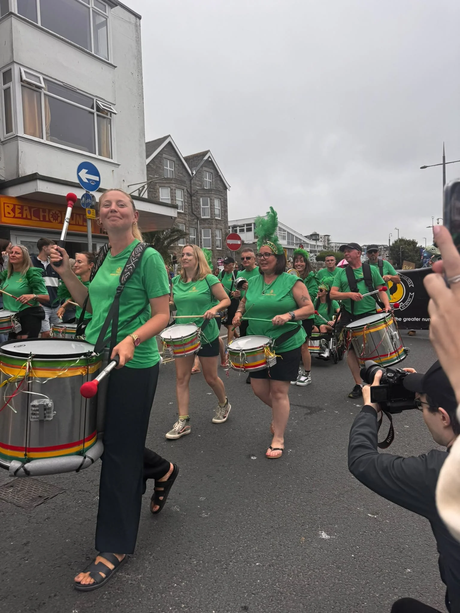 Group of women in green tops playing drums during a parade or festival on a city street with buildings and street signs in the background.