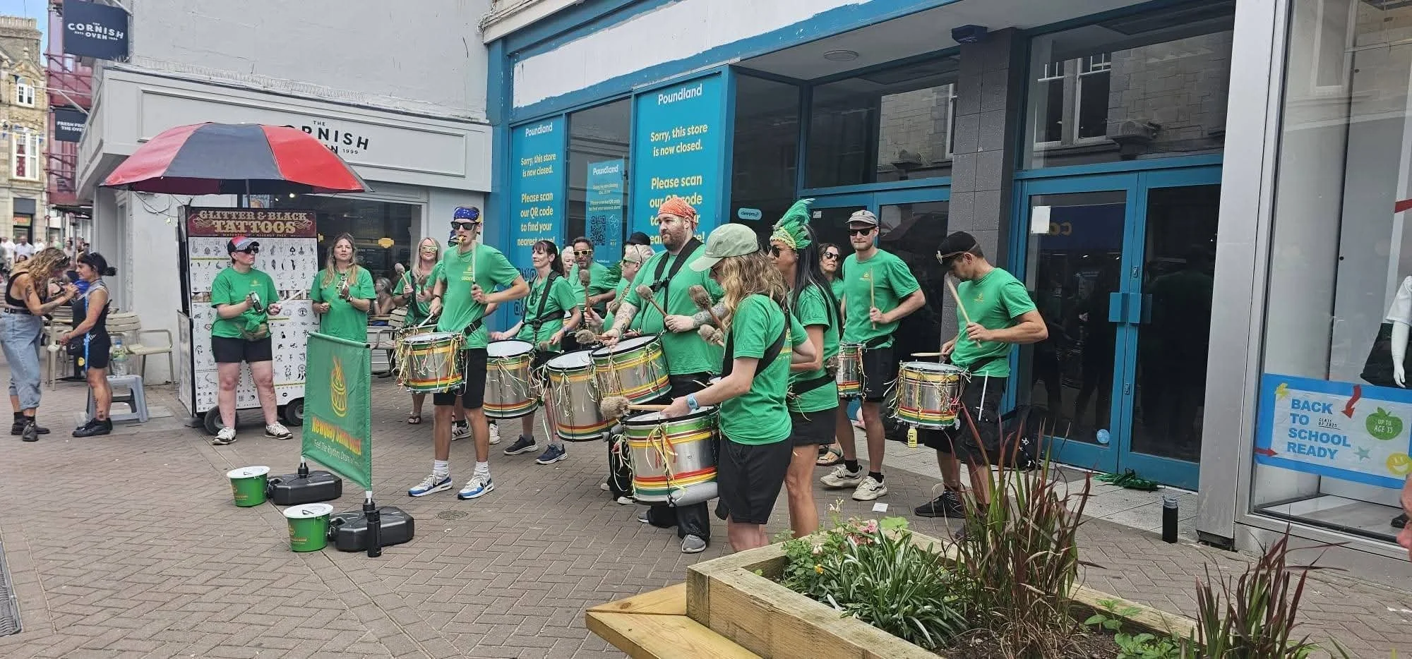 Group of street musicians playing drums and percussion outside a store with a blue Sign reading 'Poundland'. People on the sidewalk, some taking photos, and a tattoo tattoo booth nearby. Several performers wear green shirts, some with colorful headpi