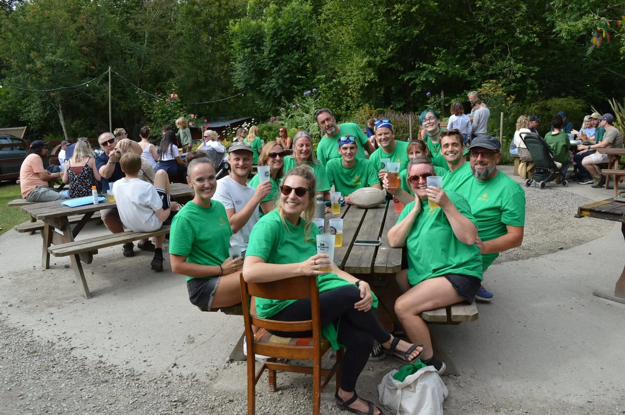 A group of people smiling and holding drinks at an outdoor gathering with picnic tables, surrounded by trees and greenery.