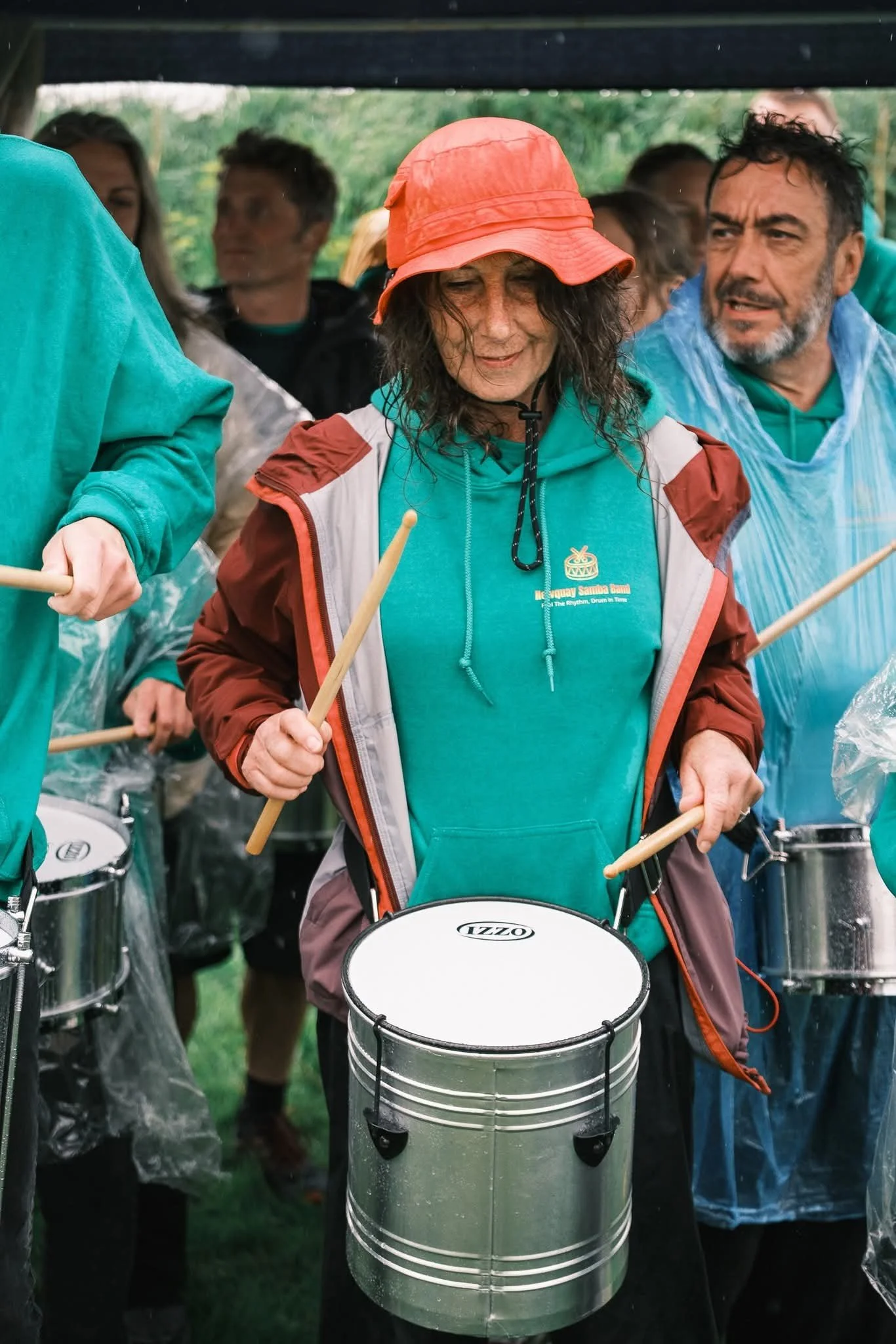 A woman wearing a red bucket hat and teal hoodie playing a small drum in a group of people, some of whom are playing drums, outdoors under a shelter.