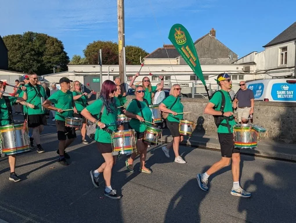 A group of people in matching green shirts and shorts marching in a parade, playing drums and carrying flags on a city street.