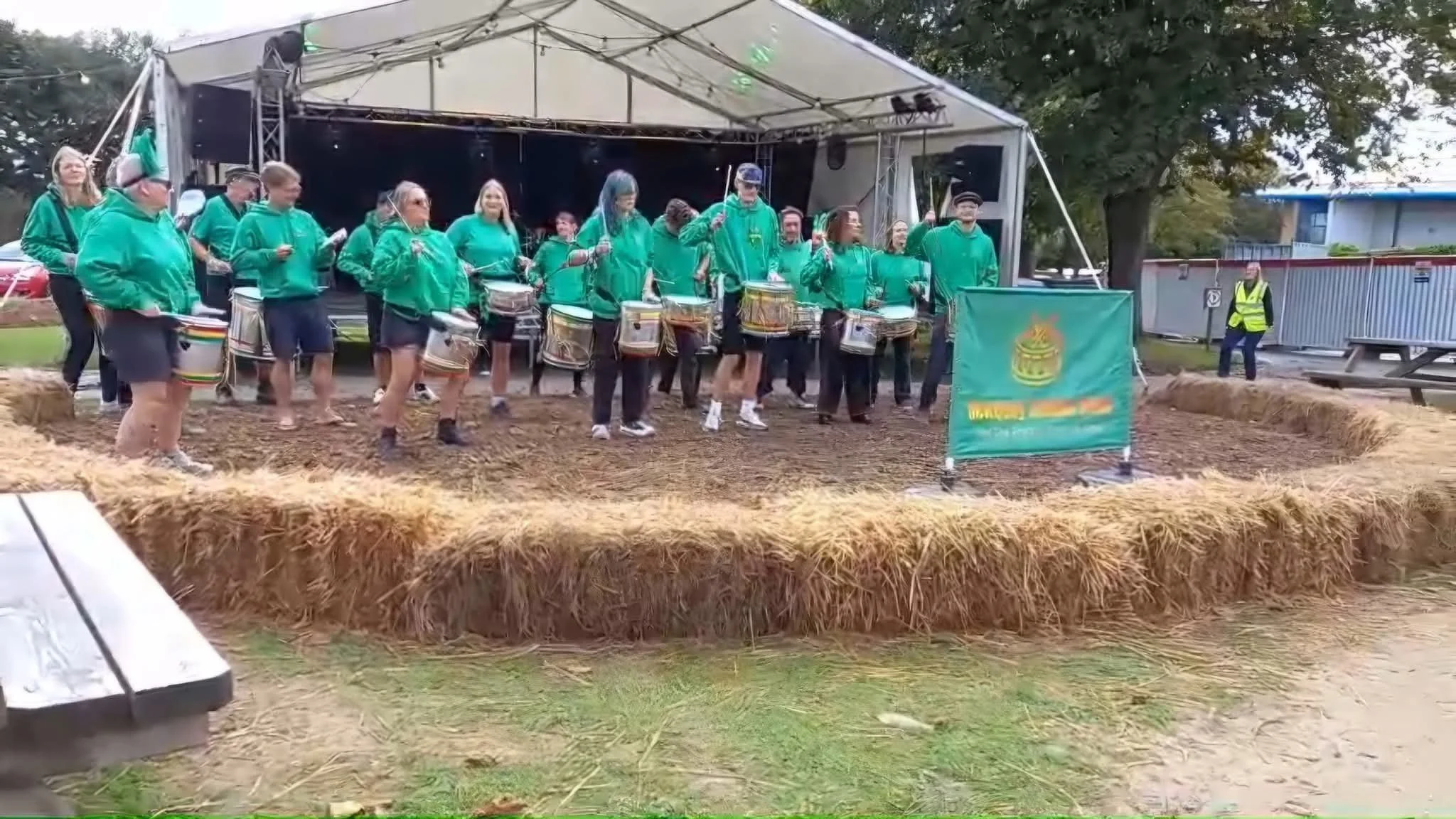 Group of people wearing green jackets and shorts, playing drums on a stage at an outdoor event with hay bales surrounding the stage.