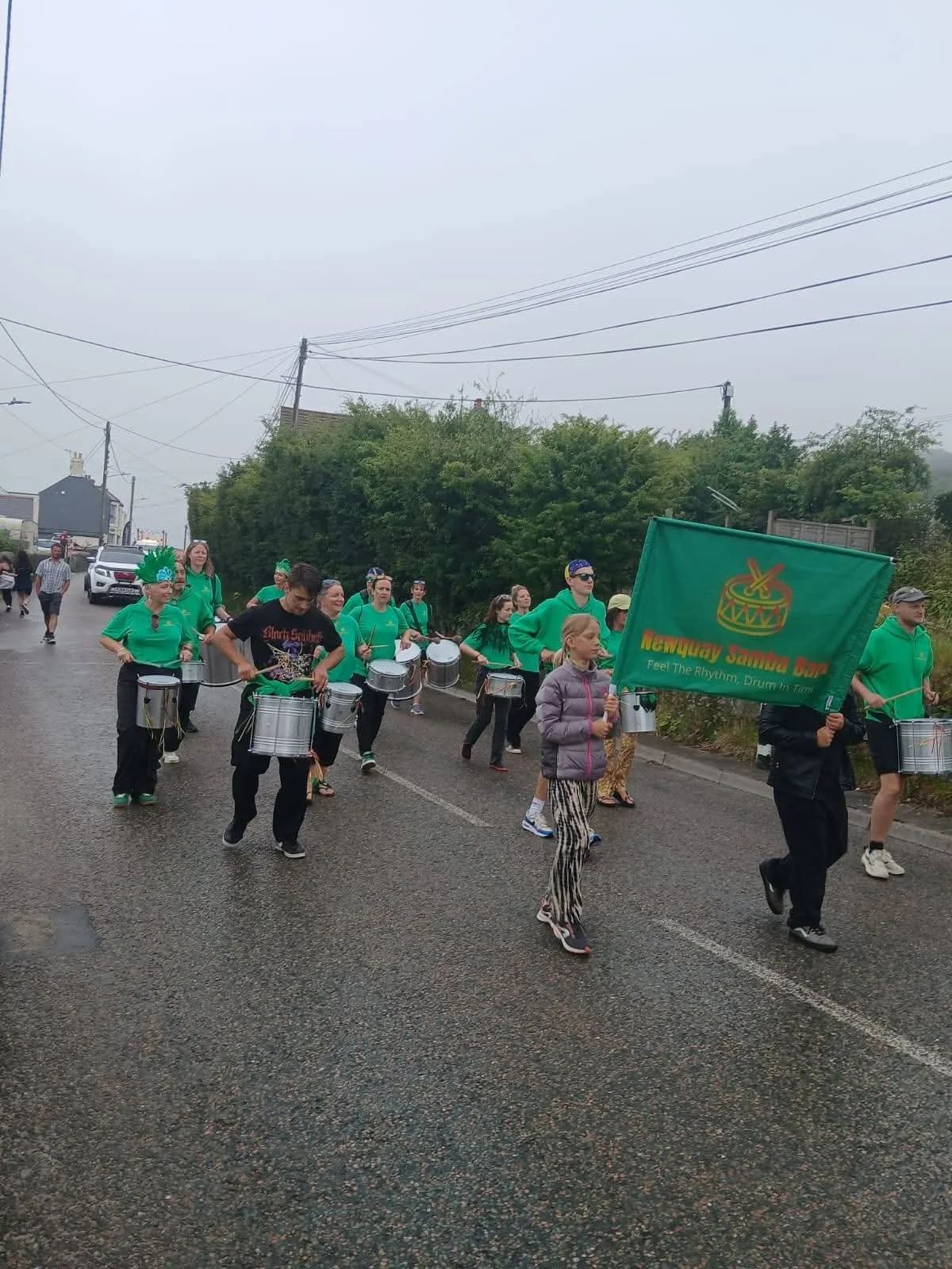 People marching in a parade with drums, some wearing green shirts and one person with a feathered headdress, and a young girl holding a green flag that says 'Newquay Samba Band'.