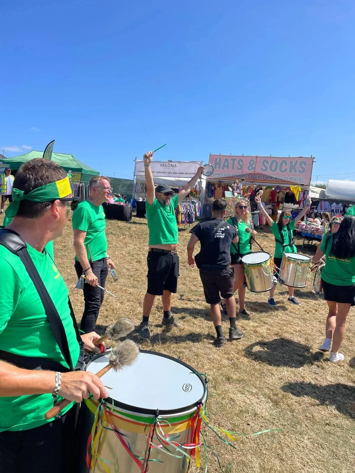 People wearing green shirts, some with headbands, playing drums and percussion instruments in an outdoor festival or fair. Vendor booths and signs are visible in the background under a clear blue sky.